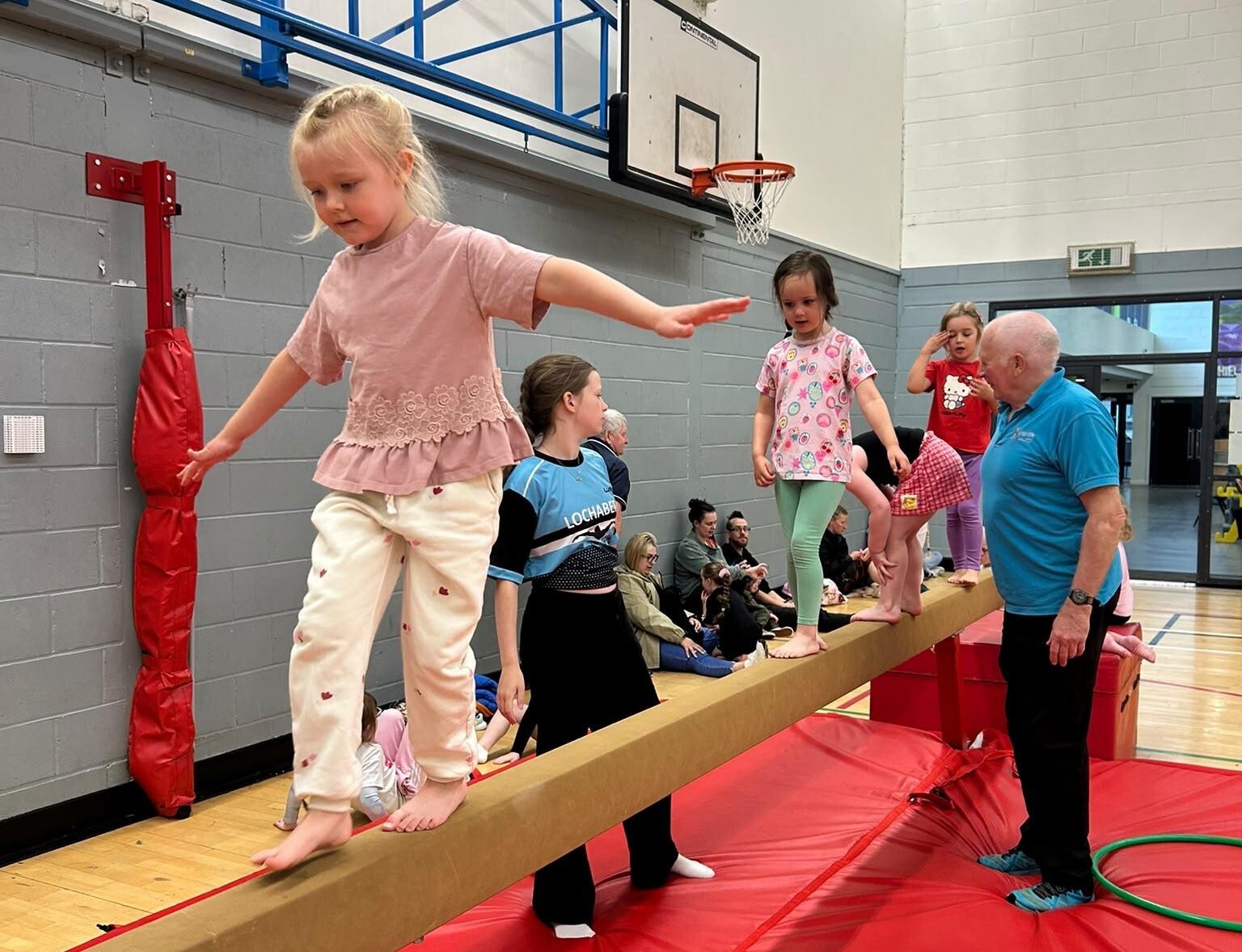 Children walk along a balance beam over red safety mats, supported by a coach and a young helper.