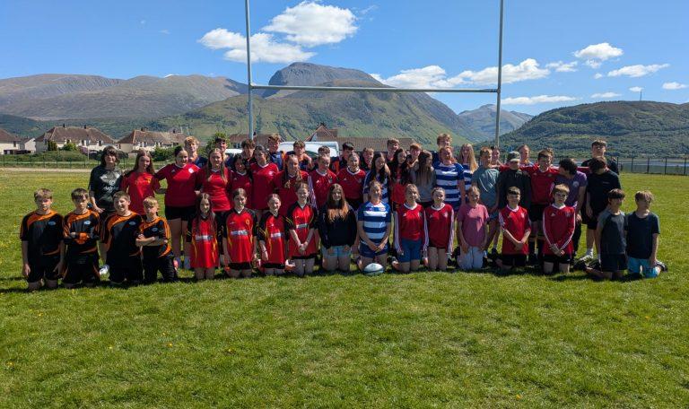 A large group of children and teenagers in sports uniforms standing on a grassy field with mountains in the background, during a rugby event in Lochaber.