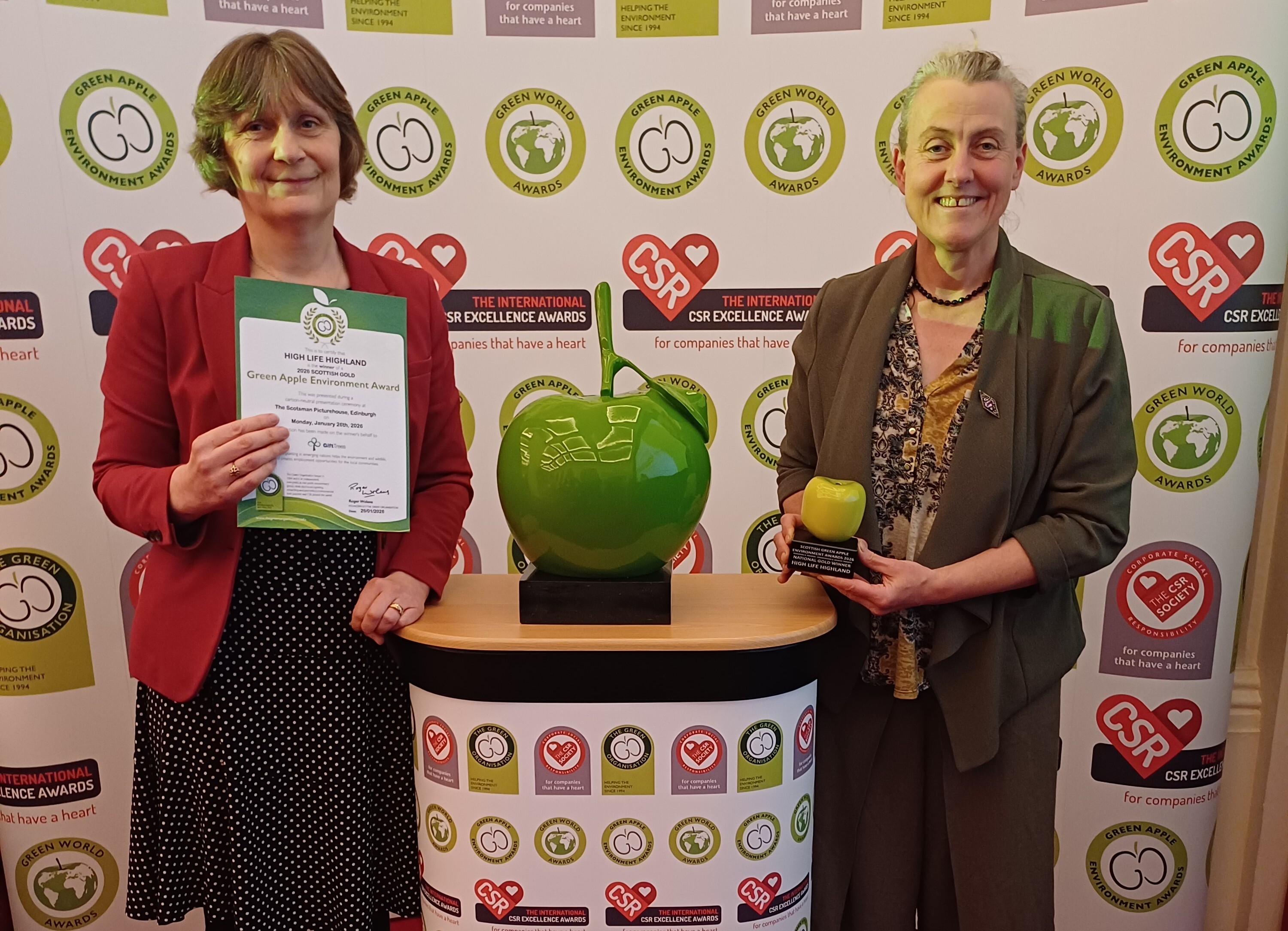 A lady in a red jacket and black dress is holding an awards certificate. On the right of the image another lady in a dark green jacket and pattern blouse is holding an award in the shape of a green apple. In between them is a high table with an oversized green apple statuette.