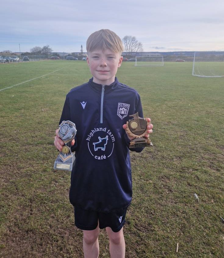 A young athlete wearing a navy sports kit stands on a grassy field holding two trophies.
