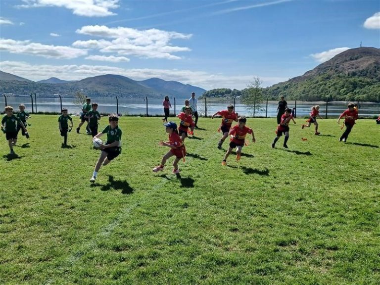 Children are playing a rugby match on a grassy field under a bright blue sky with scattered clouds. One child in a green jersey is running with the ball while several others in red jerseys chase behind.