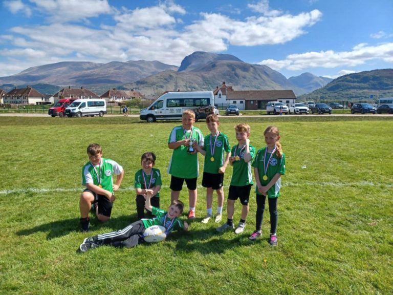 A group of six children wearing green sports jerseys with black shorts stand and sit on a grassy field, each wearing a medal around their neck. Two of the children are holding small trophies, and one child is lying on the grass in front with a rugby ball.