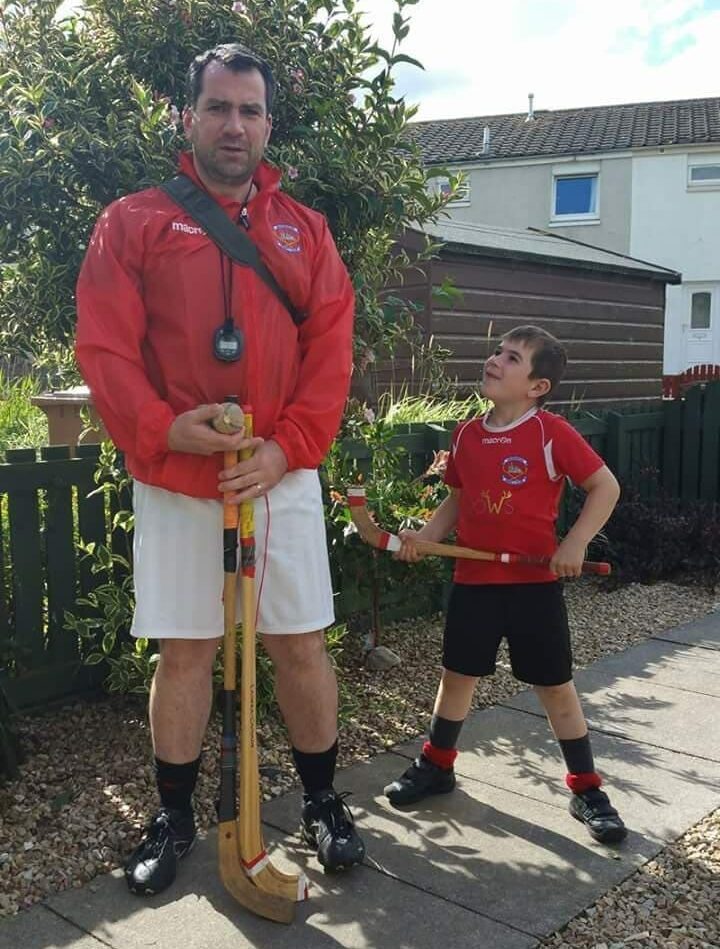 Martin Oparka and his young son standing outdoors in Inverness Shinty Club kit, each holding a caman