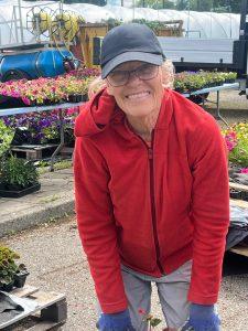 Person wearing a red jacket and black cap, bending forward while working with plants outdoors. There are trays of colorful flowers and gardening equipment in the background.