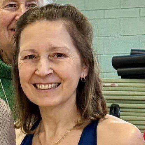 A person with shoulder-length hair smiling and wearing a sleeveless blue top, standing in front of a wall with exercise equipment visible in the background.