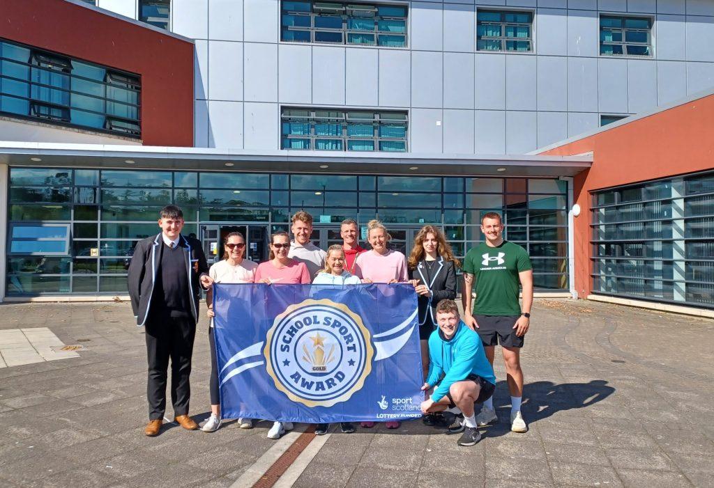 A group of ten people standing outside Millburn Academy holding a blue "School Sport Award" banner with a gold emblem.