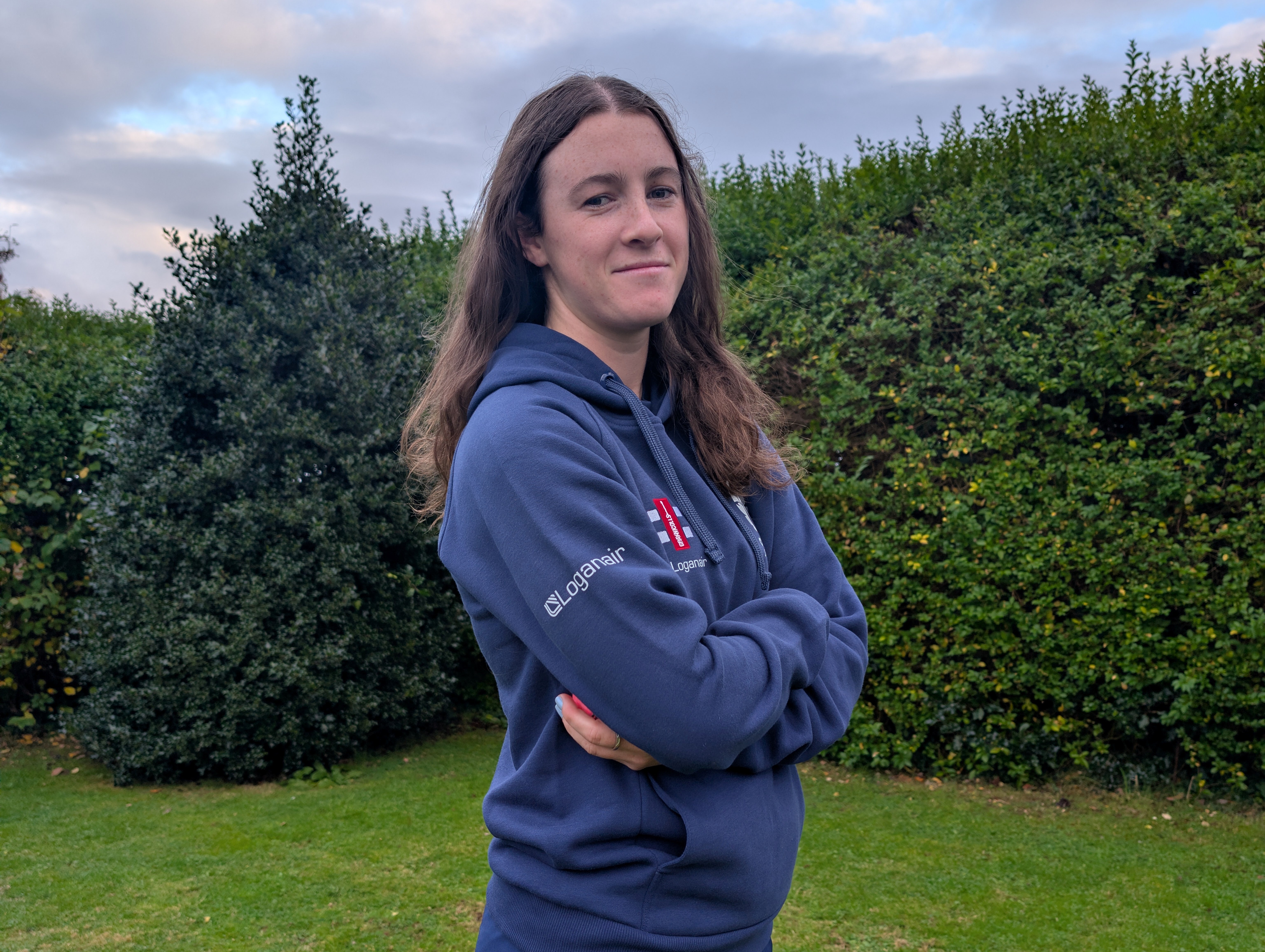 Young woman in blue jumper with long dark hair, standing in a garden. She is standing side on and looking at the camera.