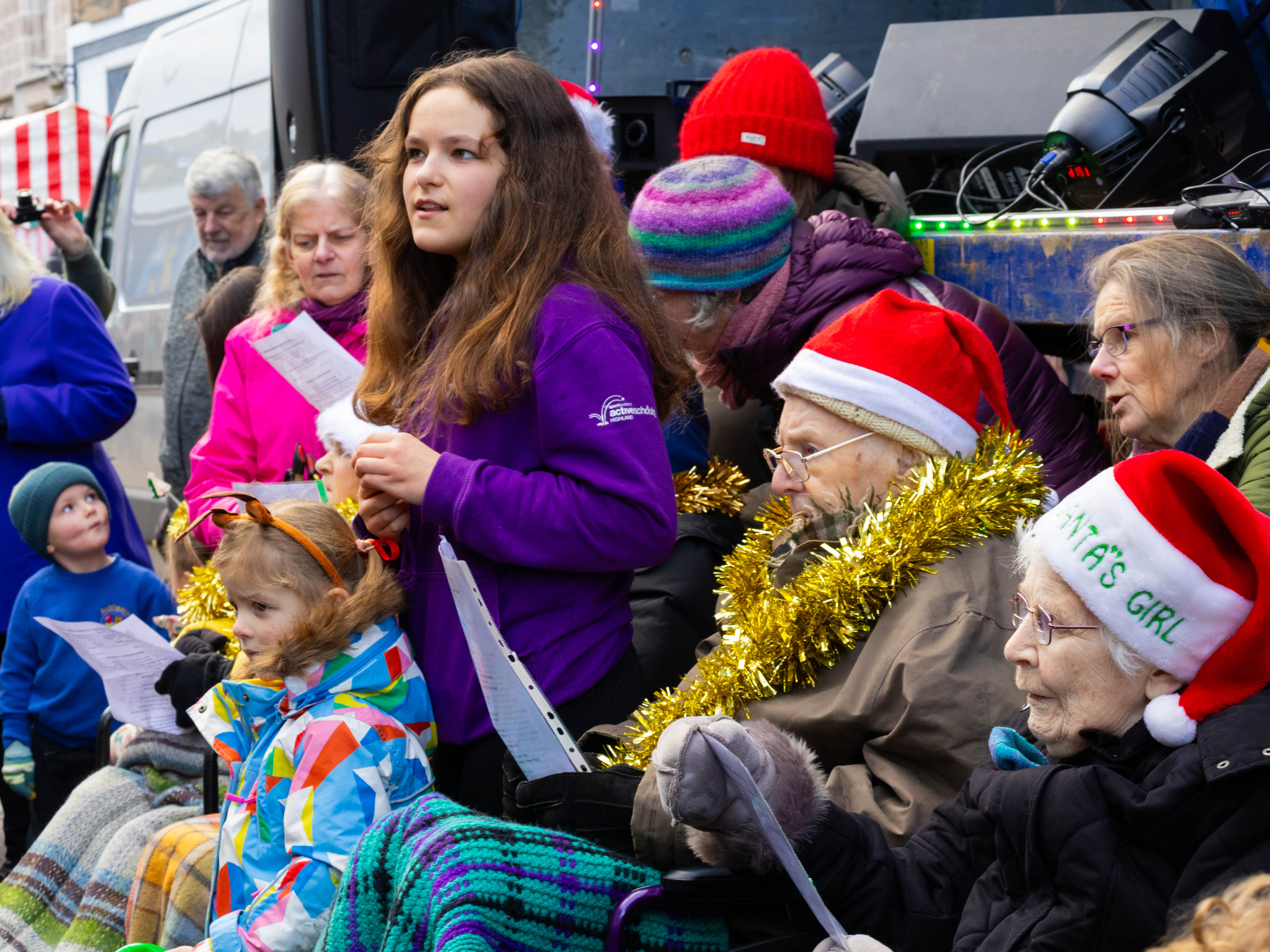 Two elderly people in wheelchairs wearing Santa hats with a young girl in a purple jumper and a small girl in front in a blue jacket. In the background a lady in a pink jacket. They are all singing.