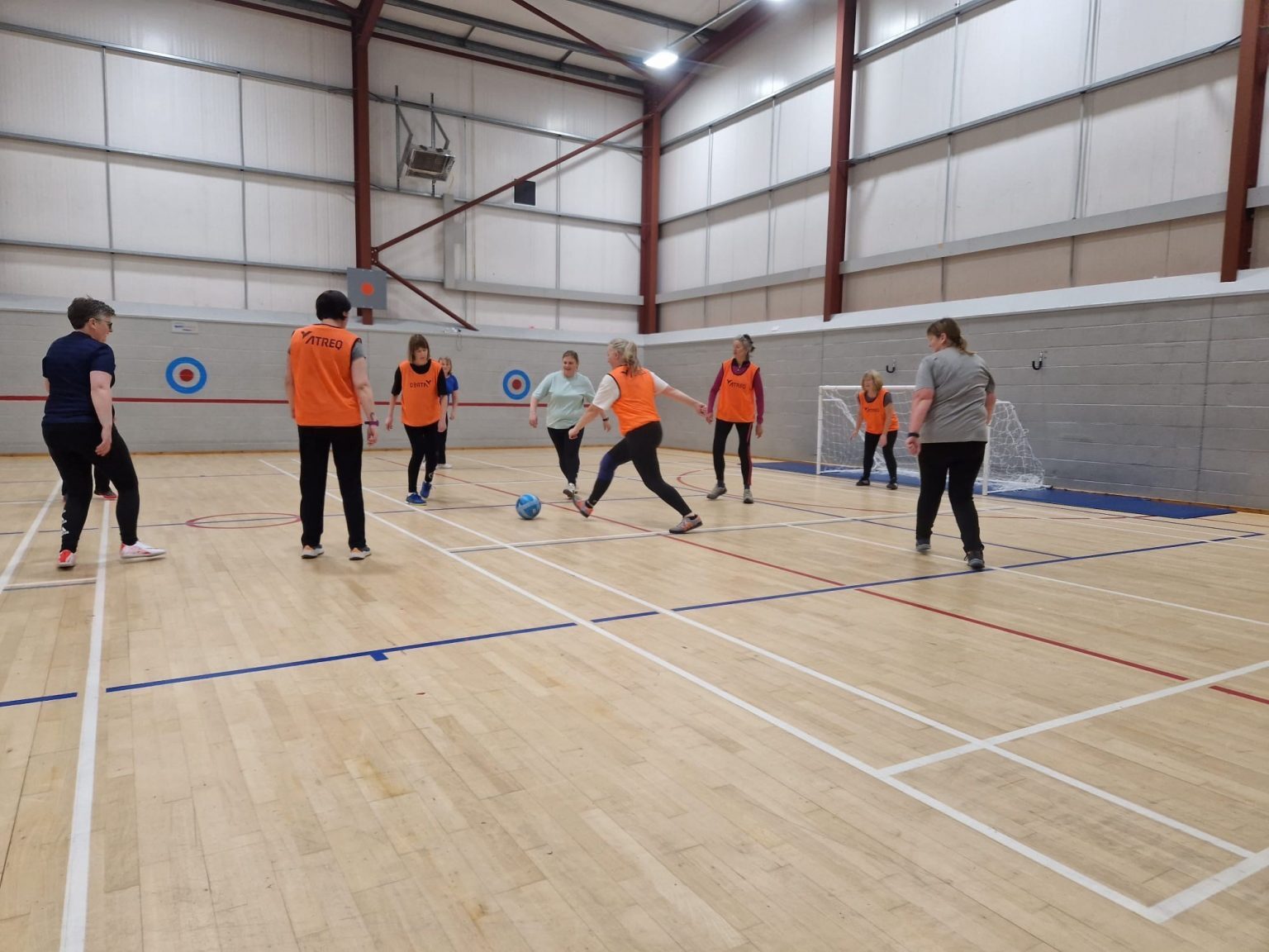 An indoor sports hall with a group of people playing a game of walking football. The wooden floor is marked with white and red court lines, and two small goals are positioned against the grey wall. Several players wear bright orange bibs, while others wear casual sports clothing. A blue football is in play at the center, with one player about to kick it while others move into position. The hall has high ceilings with metal beams and bright overhead lighting, and curling targets are visible on the walls.