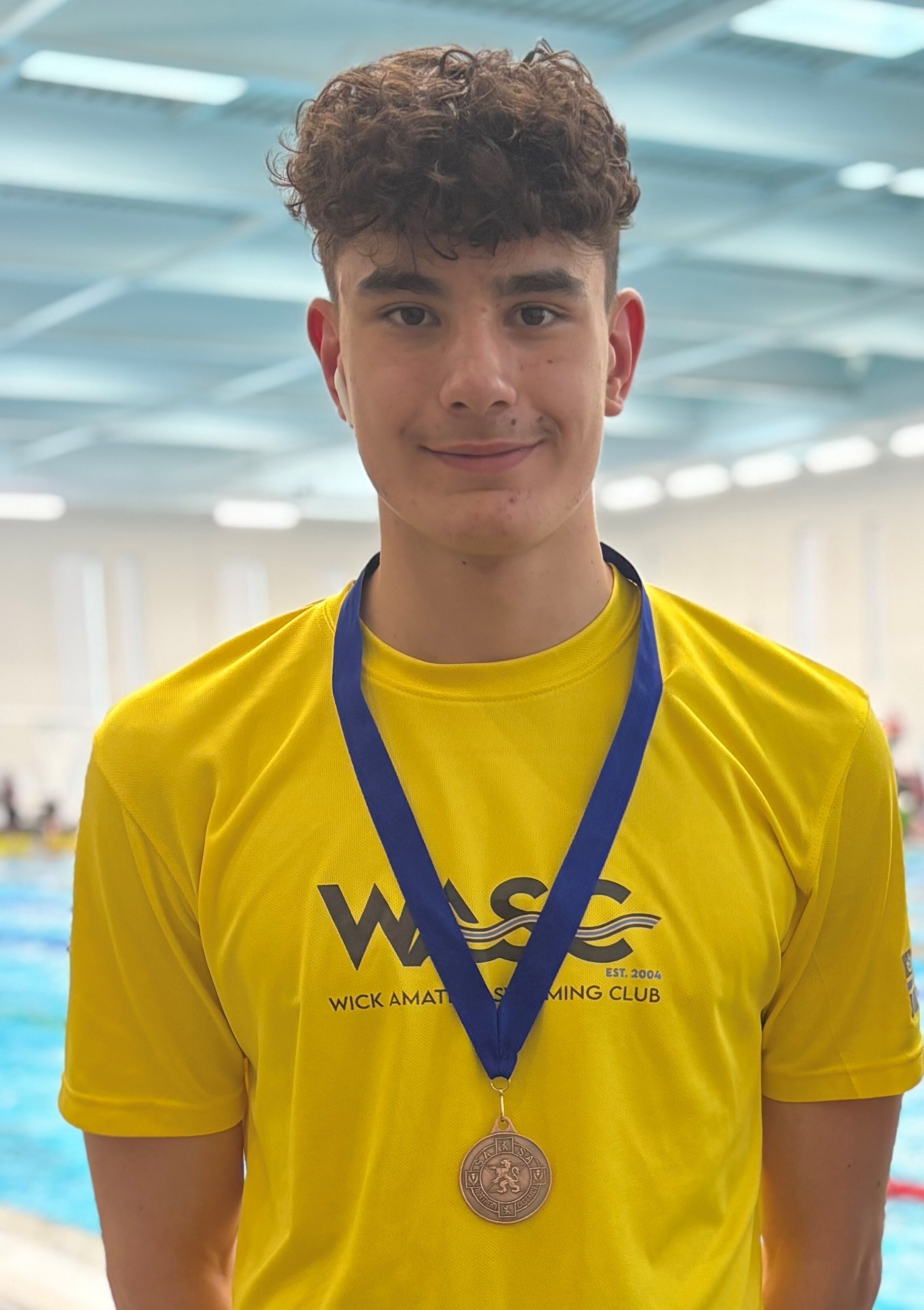 A young man with dark hair, wearing a yellow t-shirt. He is standing beside a swimming pool with a medal round his neck.