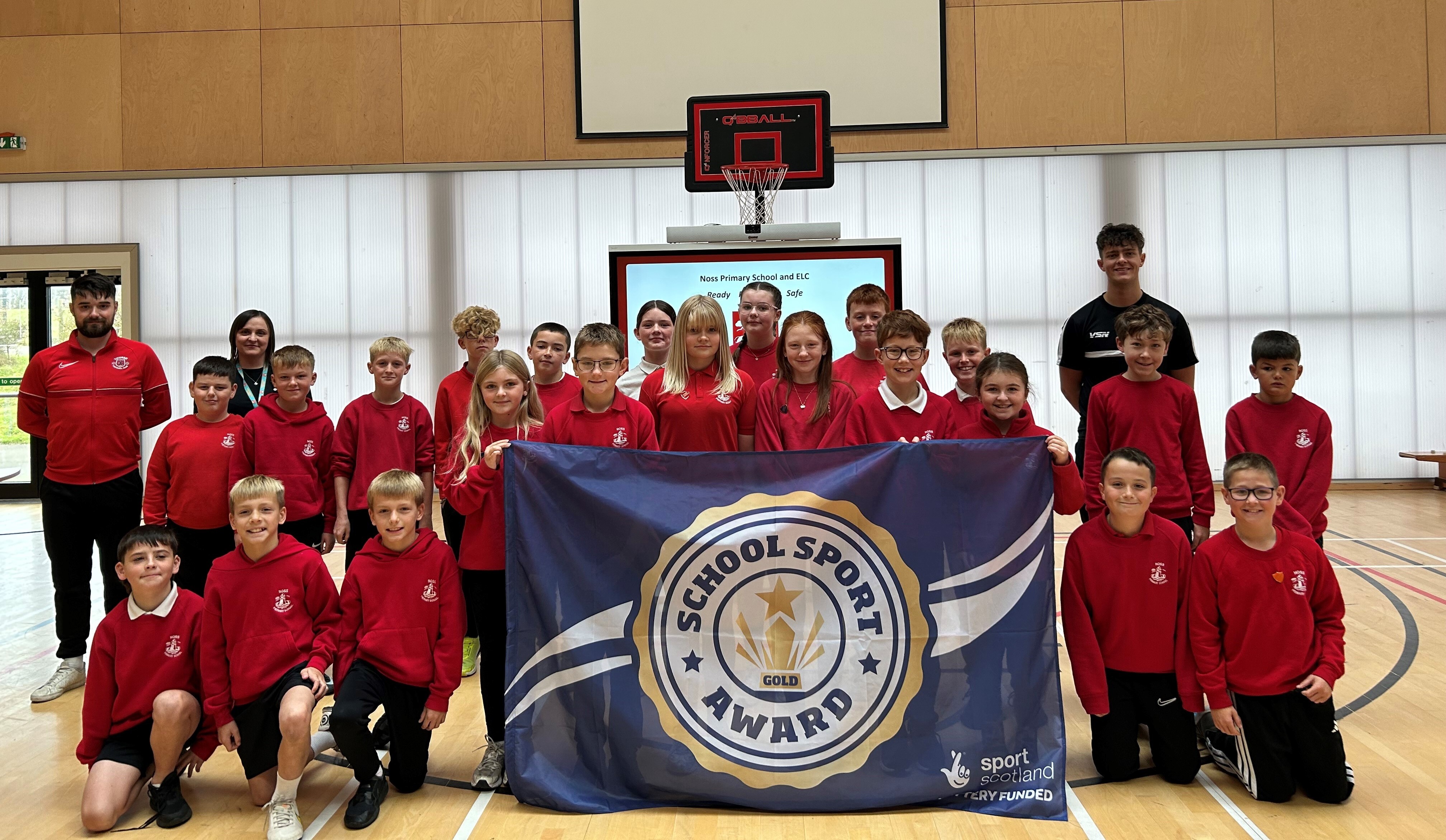 A large group of school pupils wearing red jumpers are gathered in a sports hall. They are holding up a blue and white flag which says School Sport Award