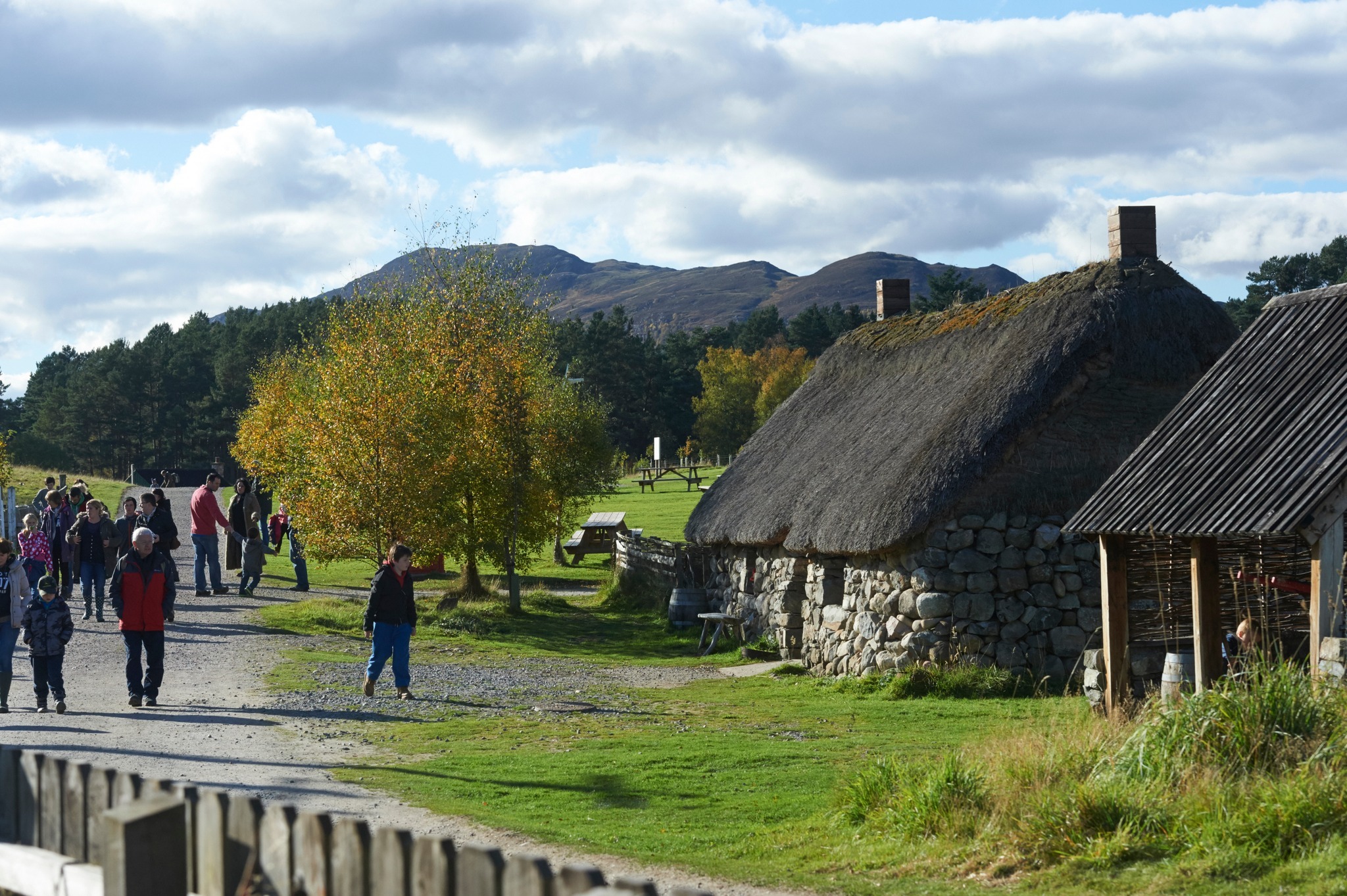 An old thatched cottage with people walking past on a dirt track. There us a tree outside the cottage and mountains in the background.