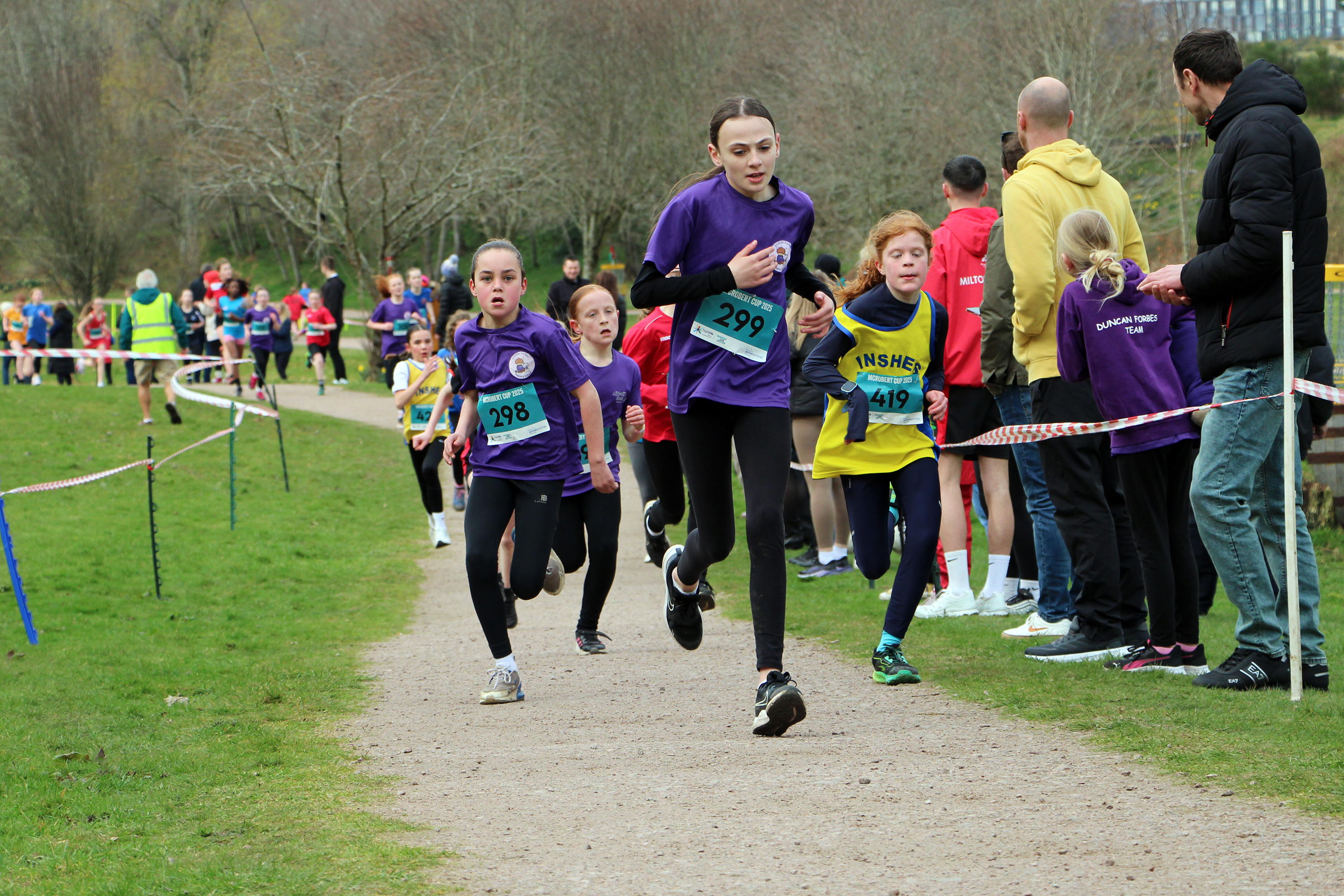 A group of female primary school pupils running a race in a park, with supporters watching close by.