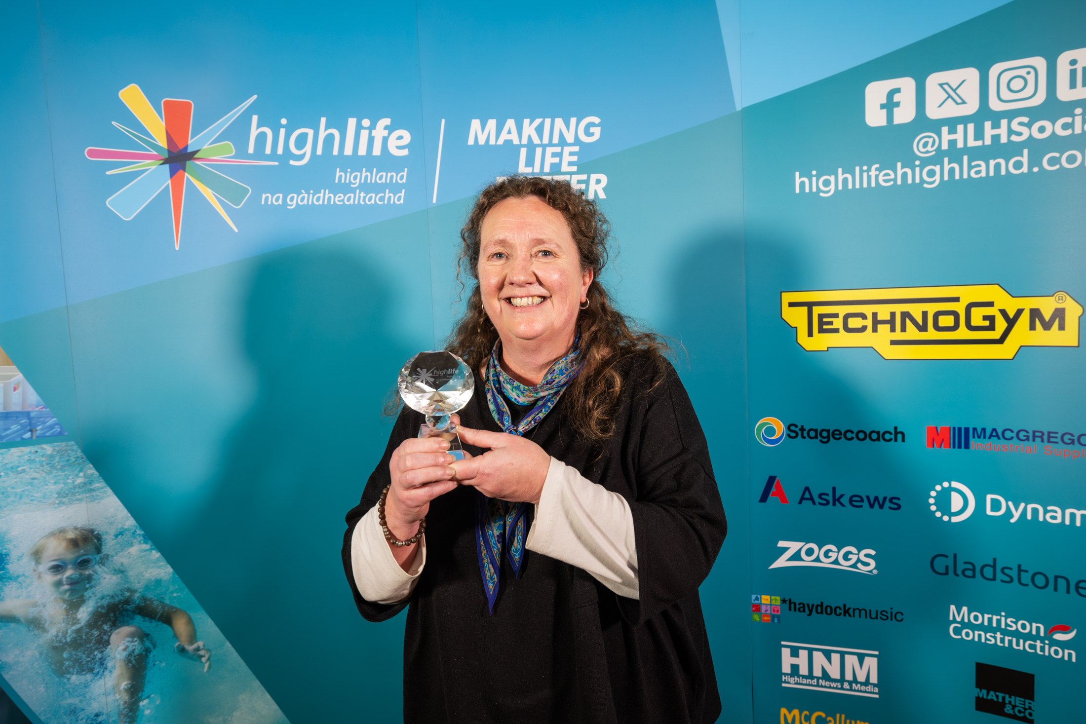 A lady with long dark hair wearing a dark dress and scarf. She is standing in front of a blue sponsorship board and holding a small glass trophy.