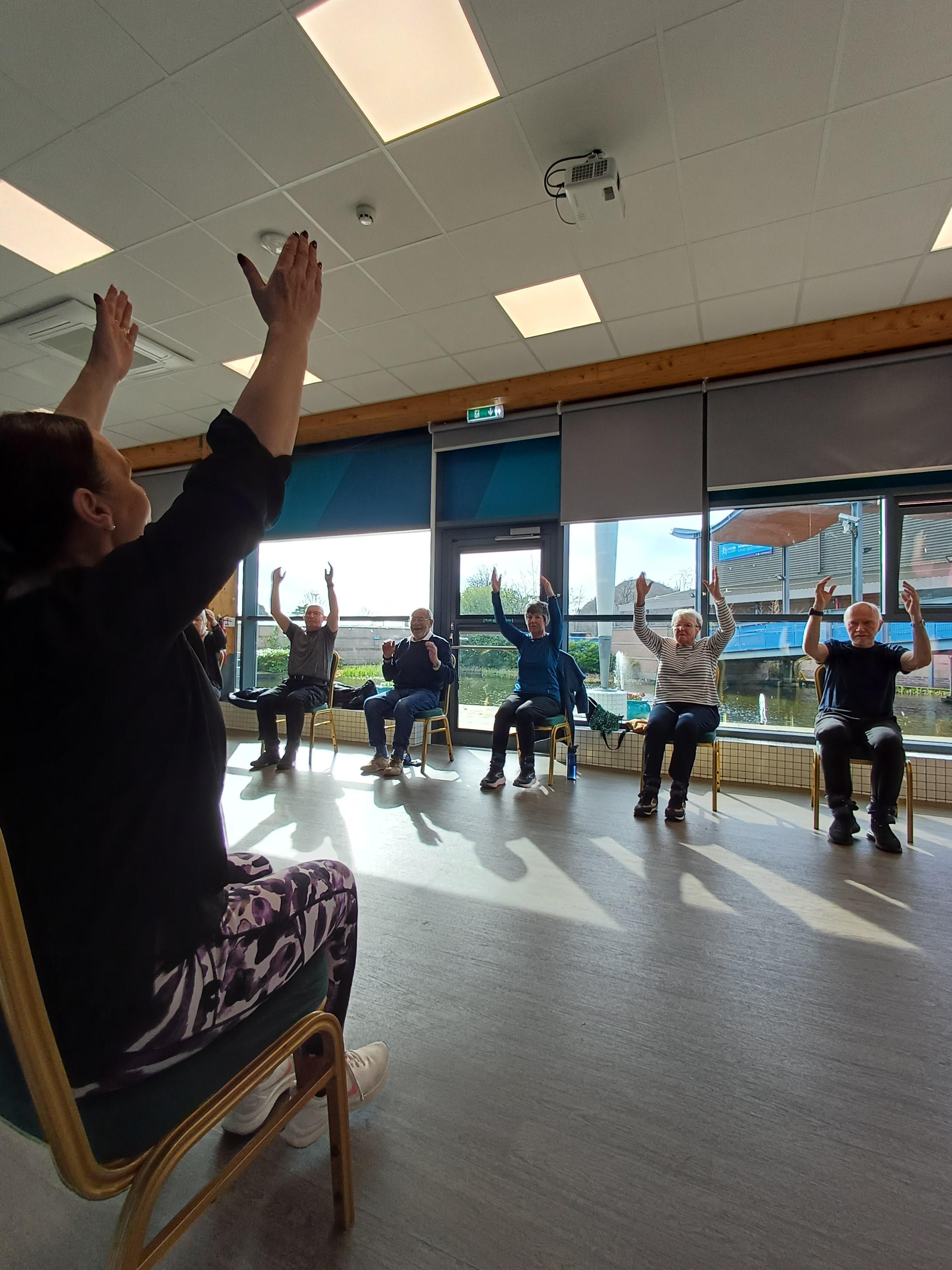 A Parkinson's exercise instructor leads a group of adults through a seated exercise class, all with arms raised.
