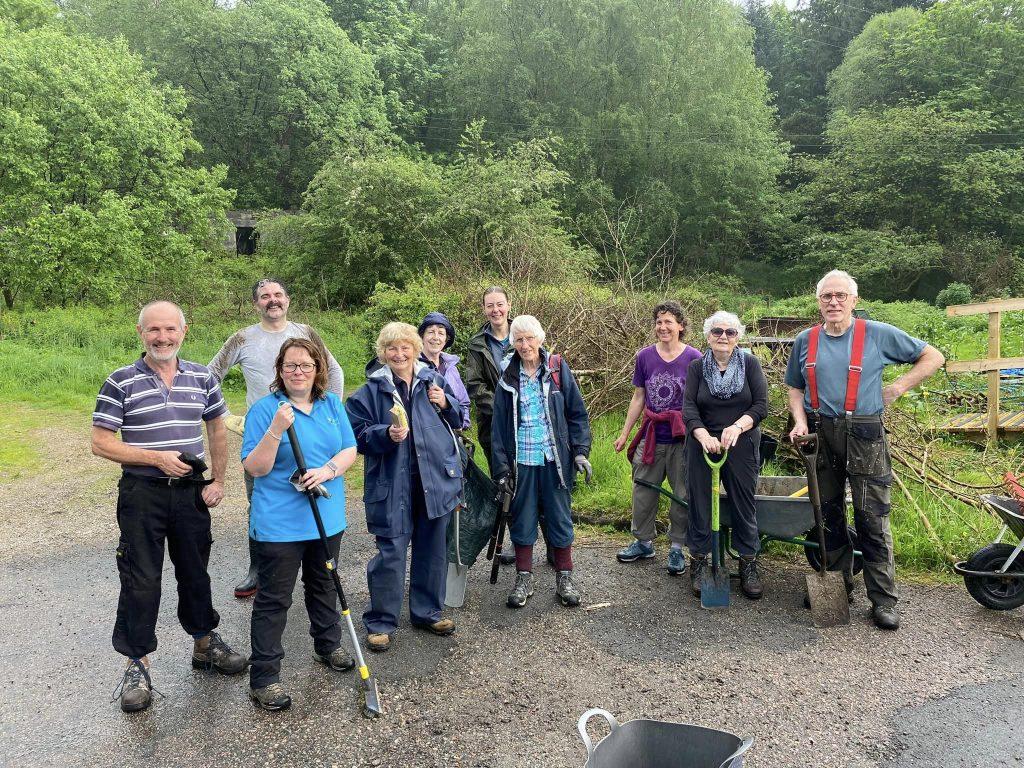 Ten volunteers standing on a gravel path in a wooded area, dressed in outdoor clothing and holding tools like shovels and rakes, with wheelbarrows and equipment nearby.