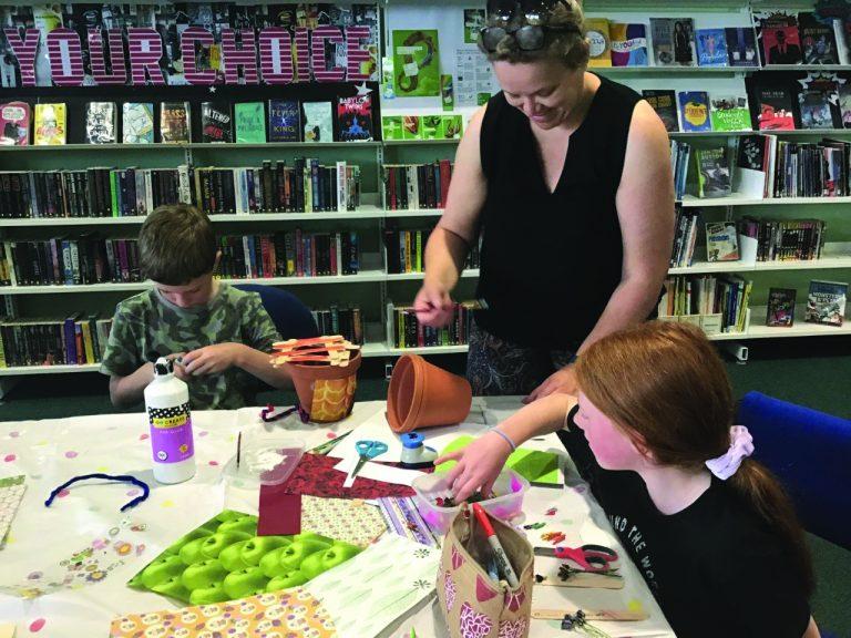 People participating in a craft activity at a library, with a table covered in materials and bookshelves in the background.
