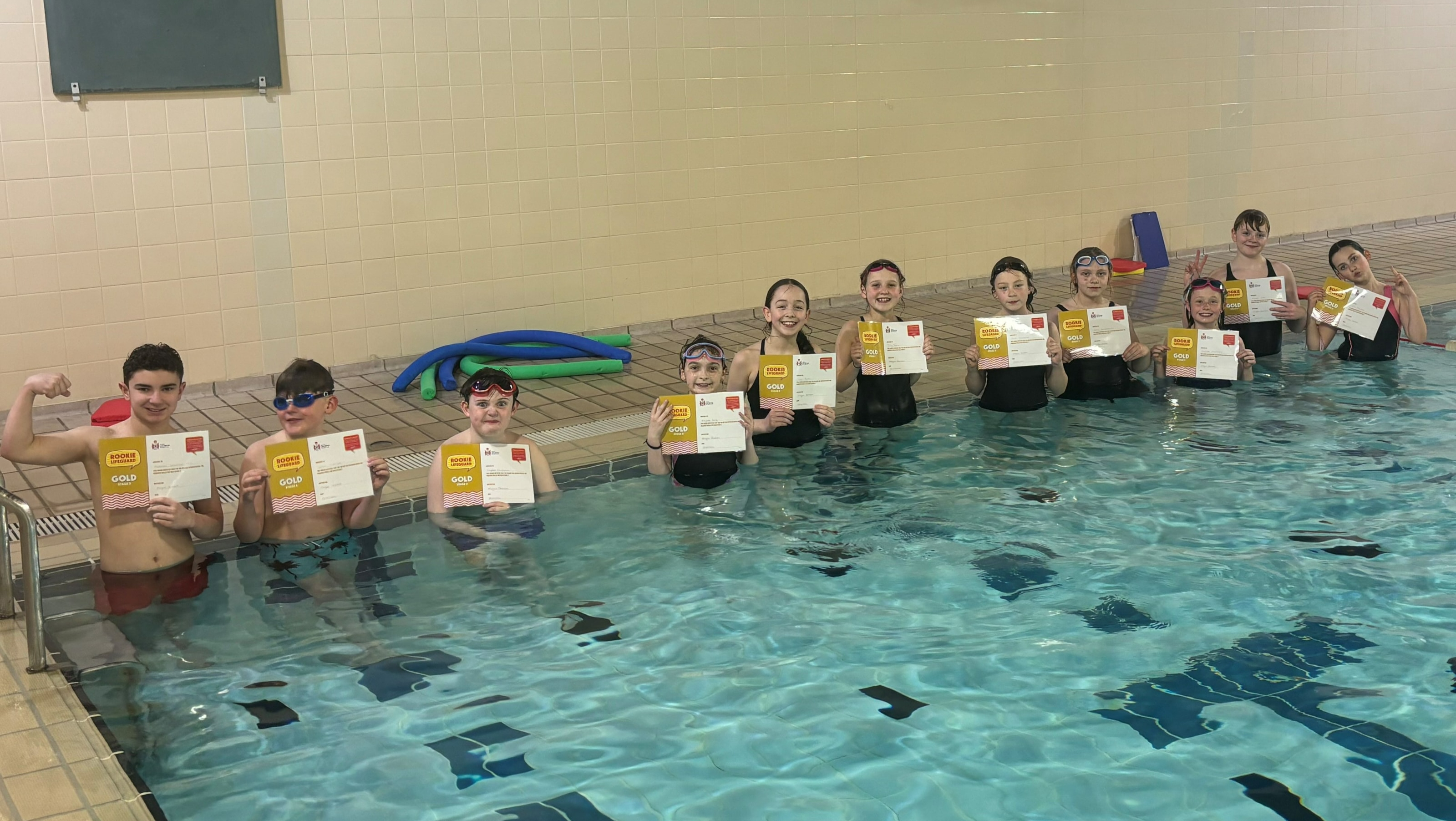 Thirteen children standing in a swimming pool holding their Gold Rookie Lifeguard certificates, smiling at the camera.