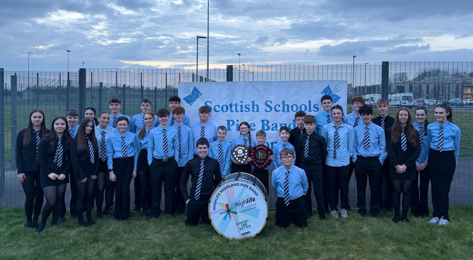 A large group of school pupils wearing dark trousers and blue shirts and ties are standing in front of a banner that reads Scottish Schools Pipe Band Championships. To the front of the group is a large bass drum and two of the younger pupils are holding competition shields.