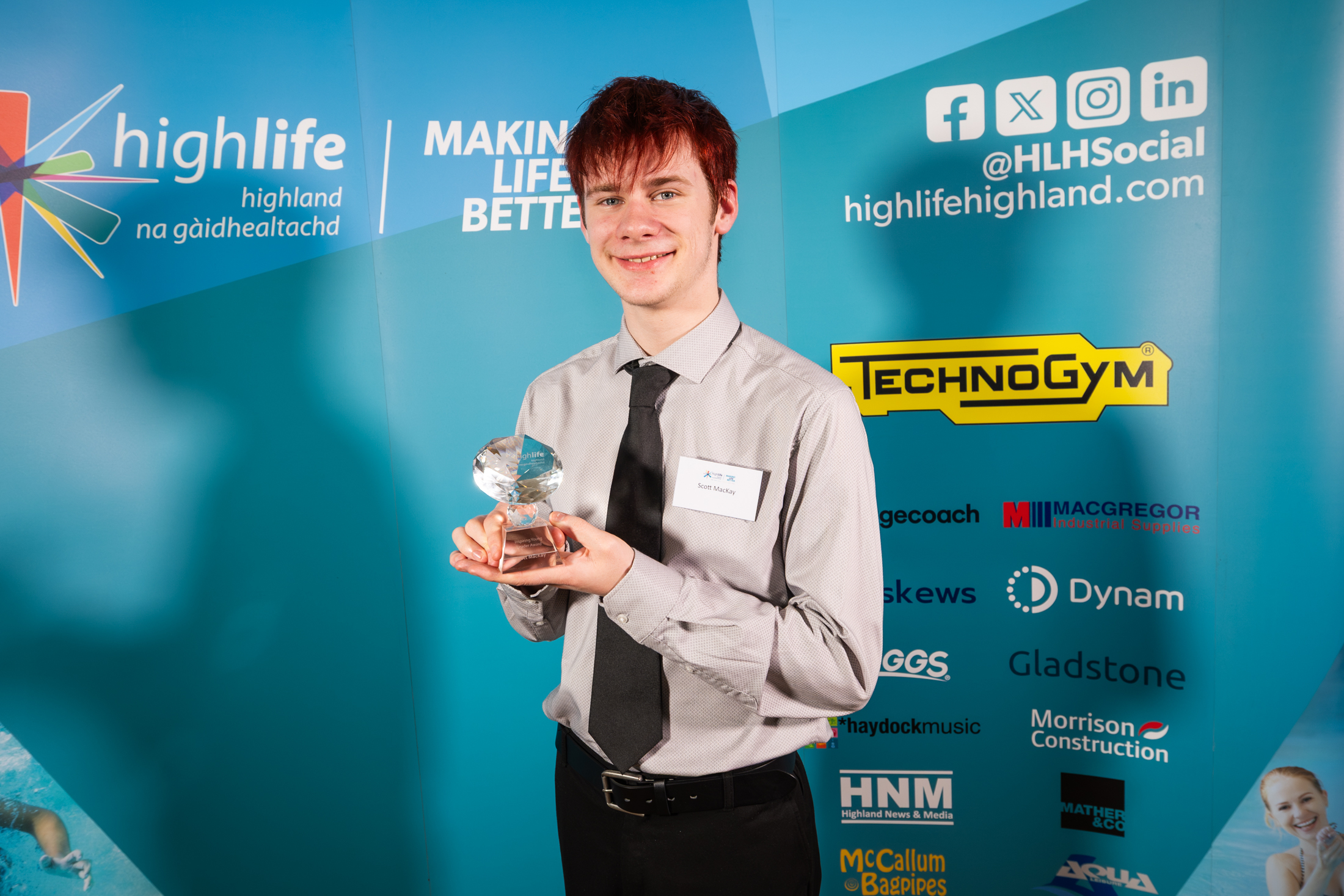 A young man in dark trousers, pale shirt and tie. He is standing in front of a blue sponsorship board and holding a small glass trophy.