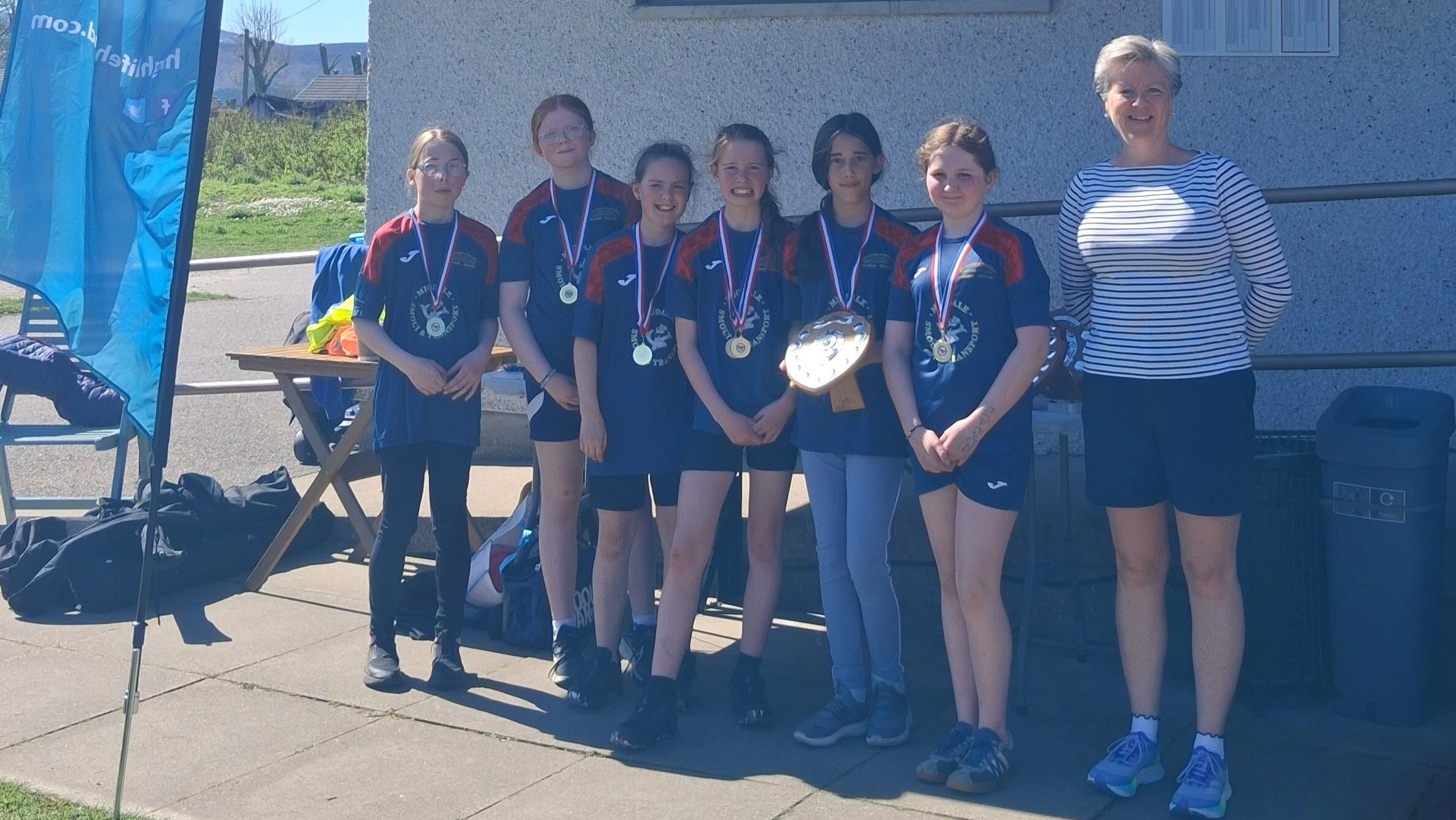 Six girls in matching navy Bonar Bridge Primary School tops holding a shield trophy and wearing medals, standing with an adult in sunshine.