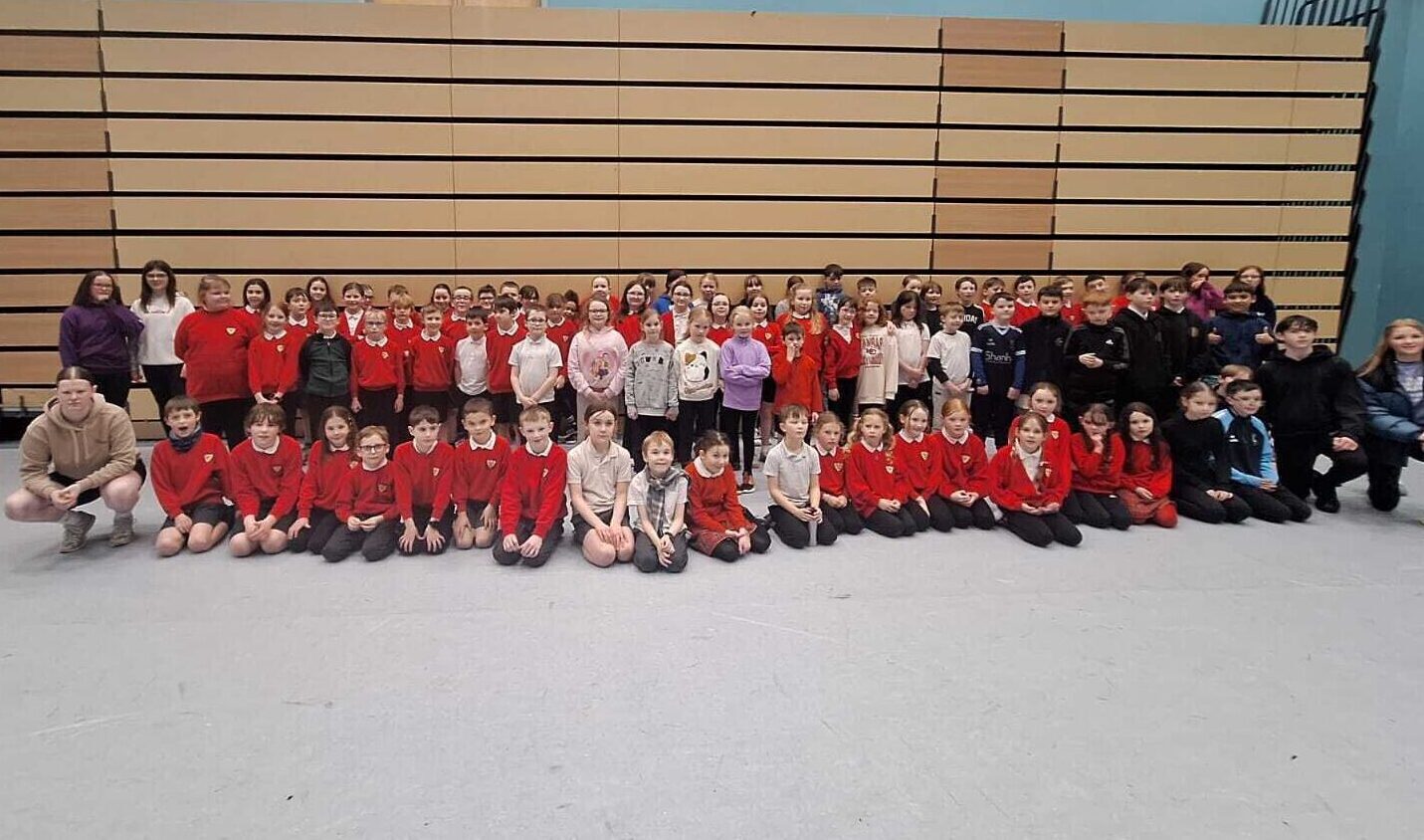 A very large group of primary school pupils, many of them wearing red jumpers. They are in a sports hall setting.