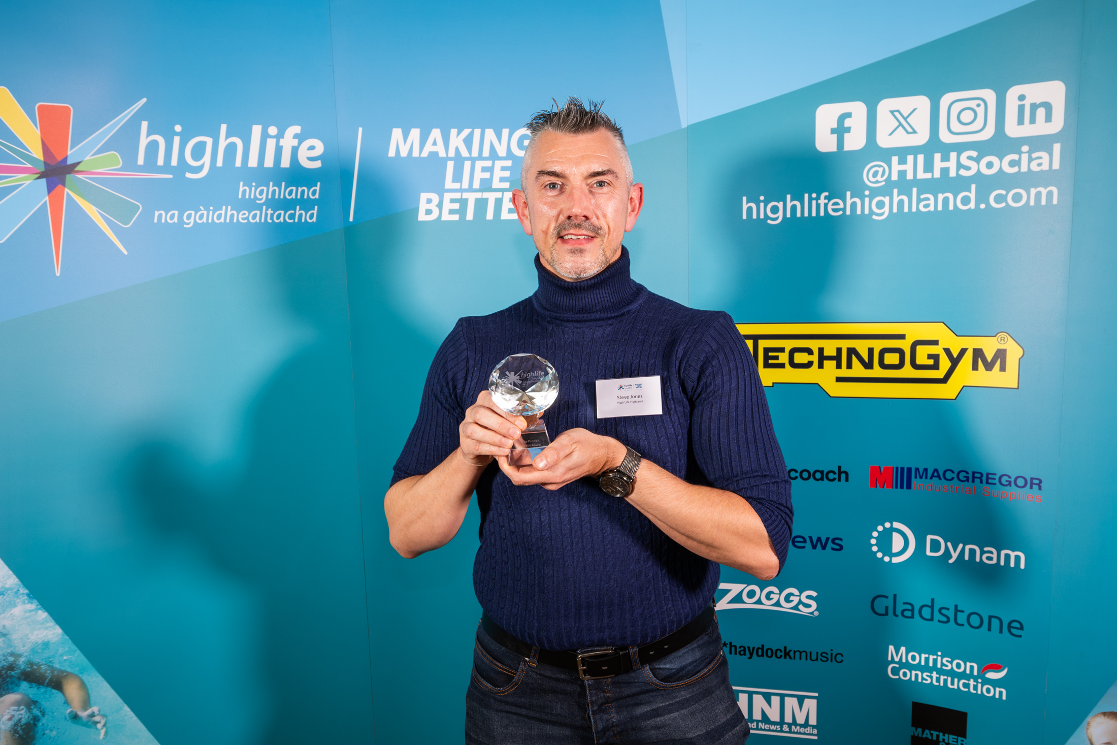 A man in dark trousers and a dark polo neck jumper. He is standing in front of a blue sponsorship board and holding a small glass trophy.