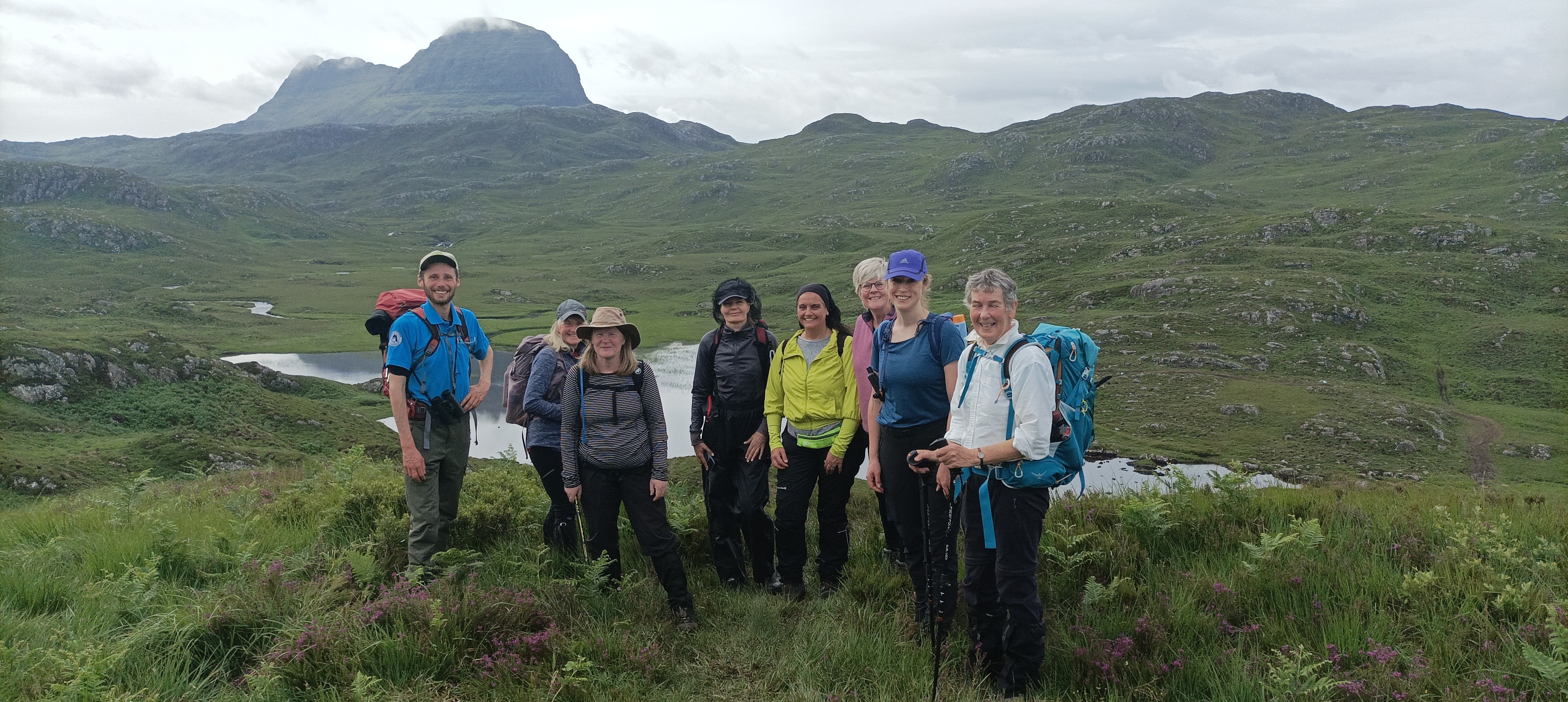 7 walkers with their guide Jack on the far left in a blue top, hat and backpack. They are standing on open moorland with Suilven in the background.