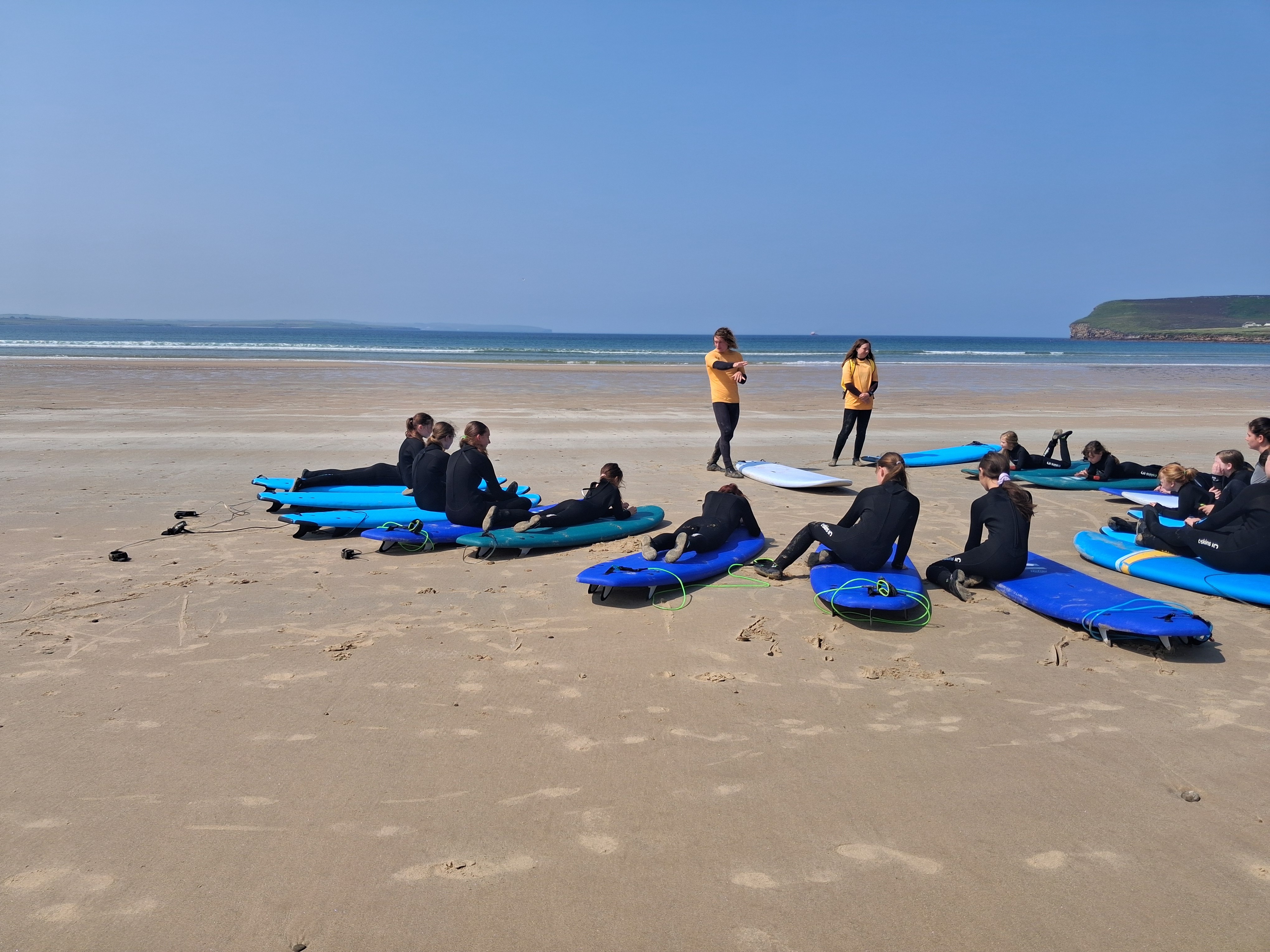 Young people in wetsuits lying on surfboards on a sandy beach while two instructors demonstrate