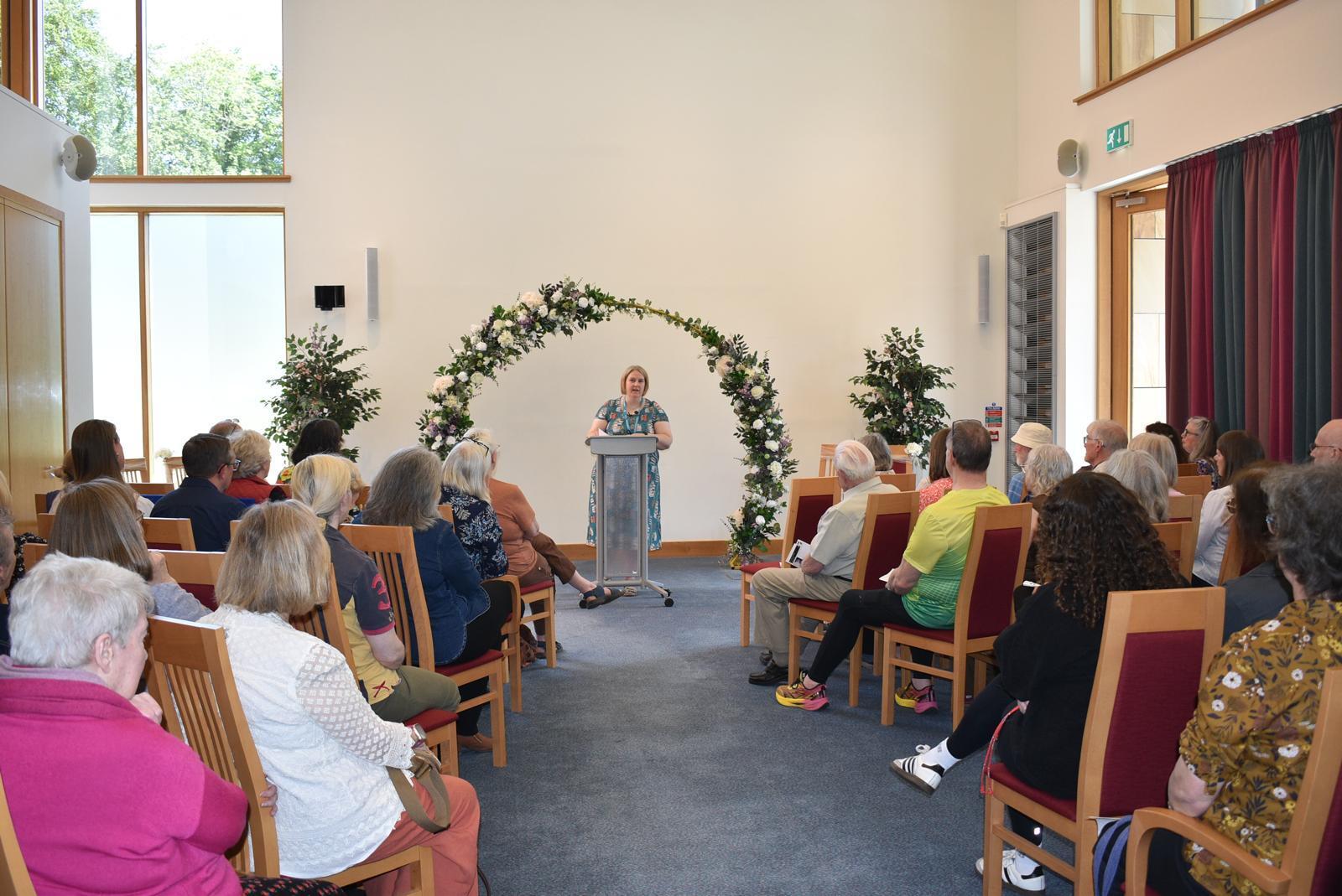 A speaker addresses a full audience at a lectern decorated with a floral arch