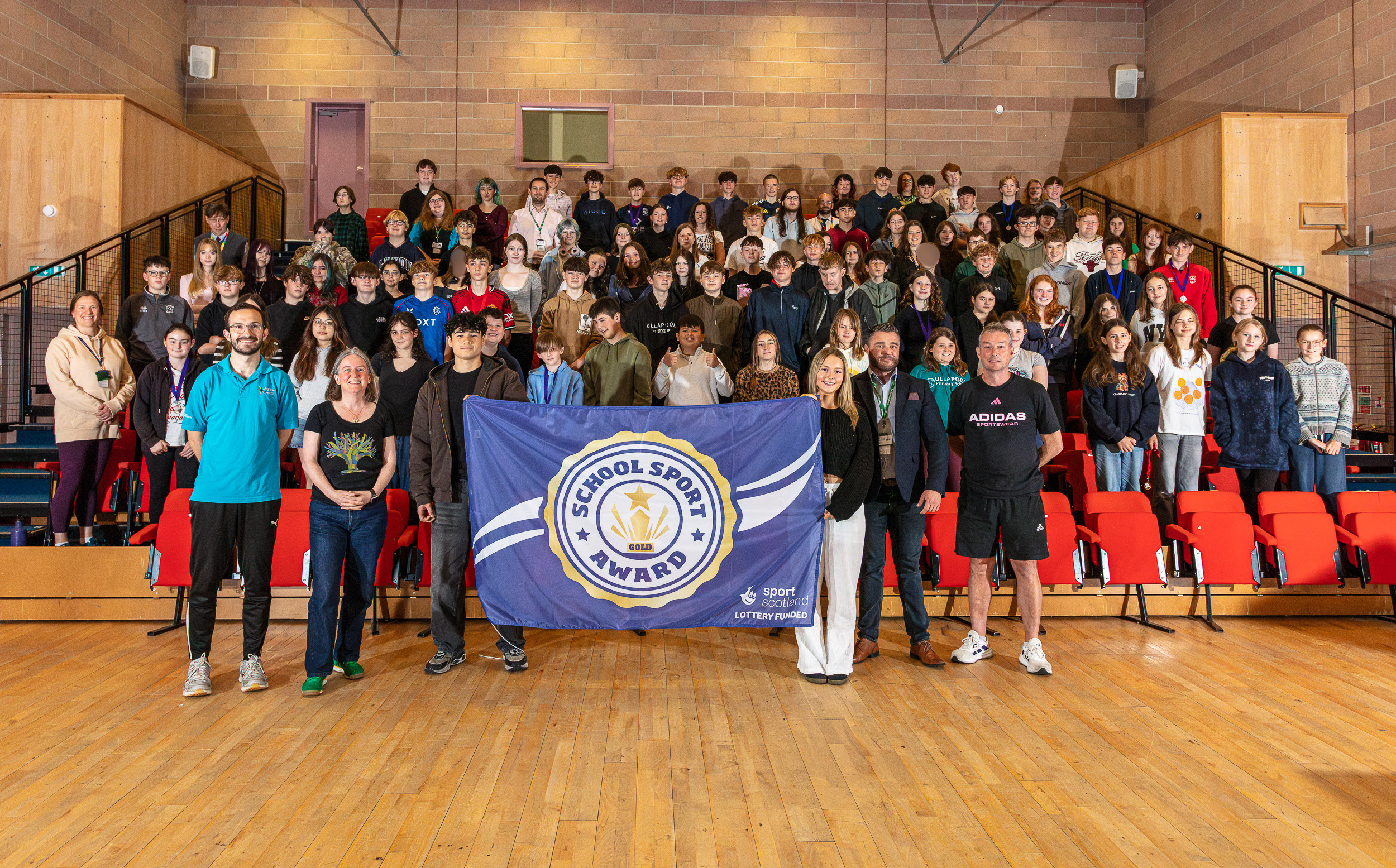 A large group of pupils and staff are gathered in an assembly hall. Standing in the front row are from left to right, Ben Bruce of High Life Highland, Maree Todd MSP, pupil Yolie De Gracia Macneilage who is holding the right corner of a blue sportscotland award flag, pupil Amy Martin who is holding the left corner of the flag. To her left are Dr Lee Coutts and David Lees, Ullapool High School staff.