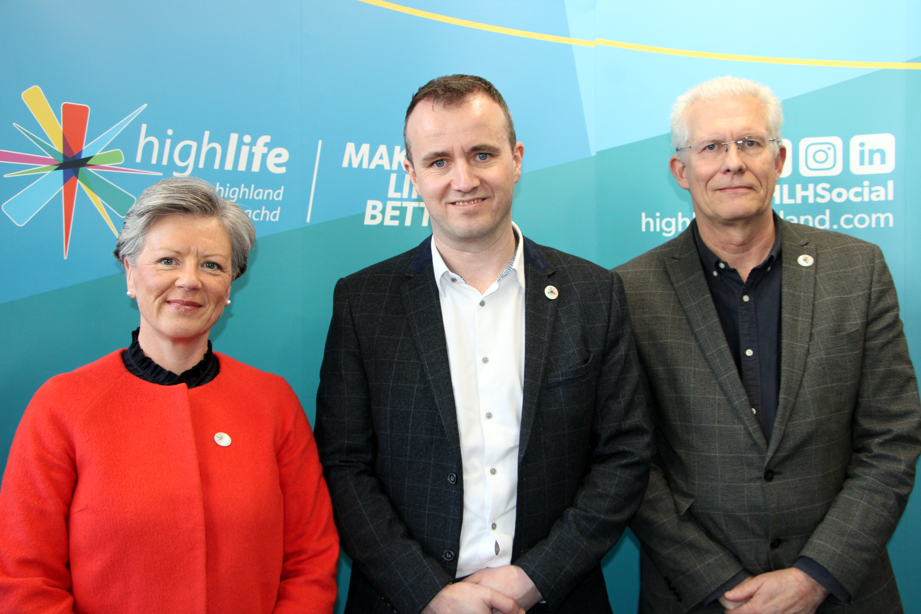 A woman in a red jacket on the left, to her right is a man in a black jacket and white shirt, to his left is an older man in a dark jacket and shirt. They are standing in front of a blue back drop.