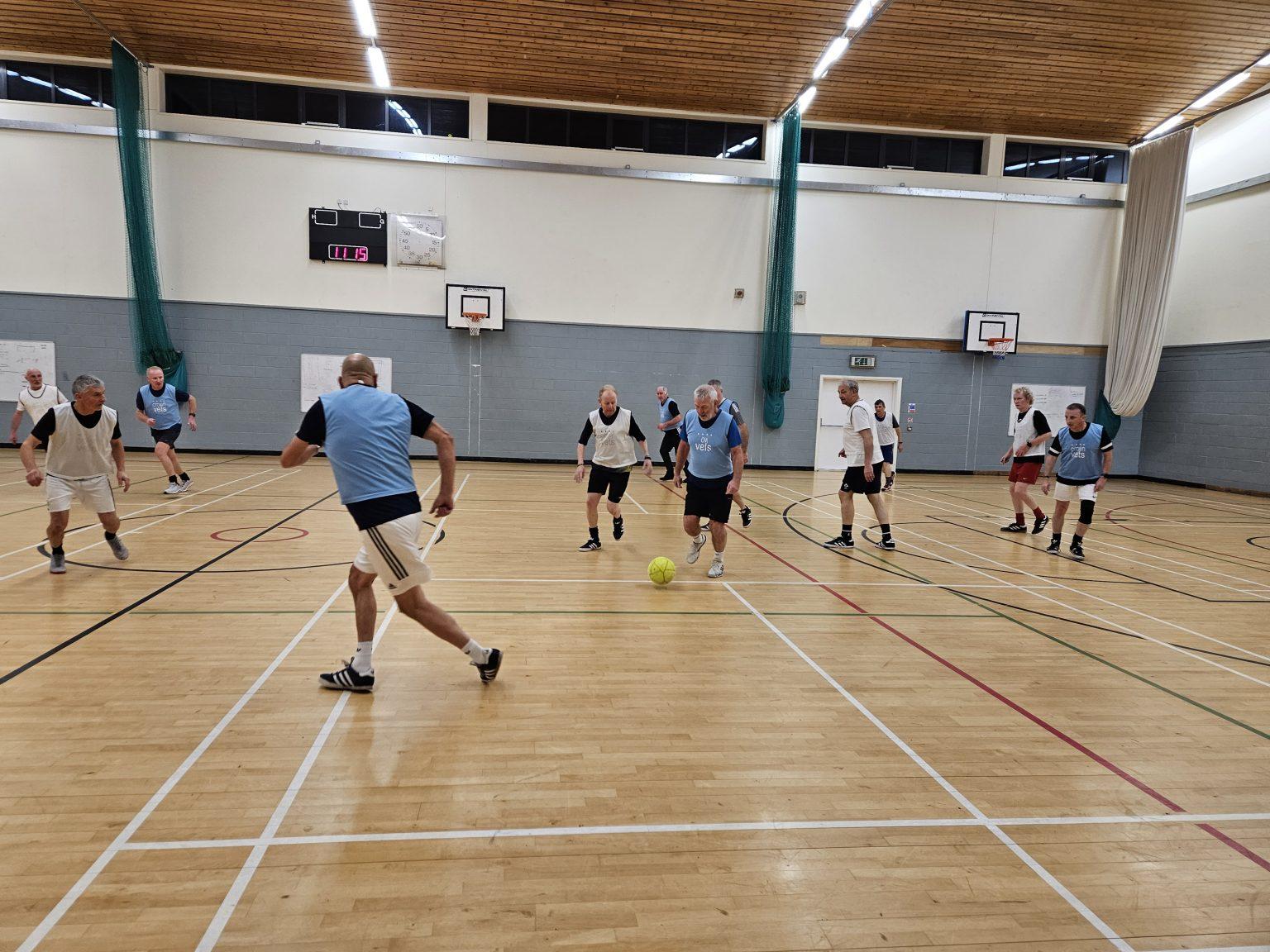 Indoor sports hall with wooden flooring where a group of older adults are playing walking football. Players are wearing blue and white bibs and moving towards a yellow ball.