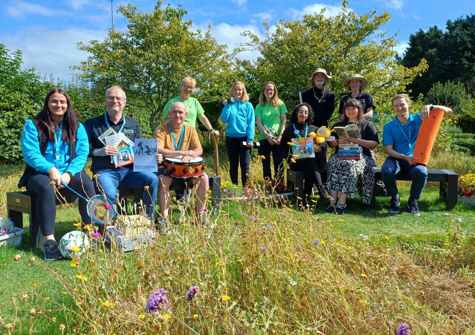 A group of eleven people in a garden with wildflowers, holding books, sports equipment, and musical instruments, with trees and a blue sky in the background.