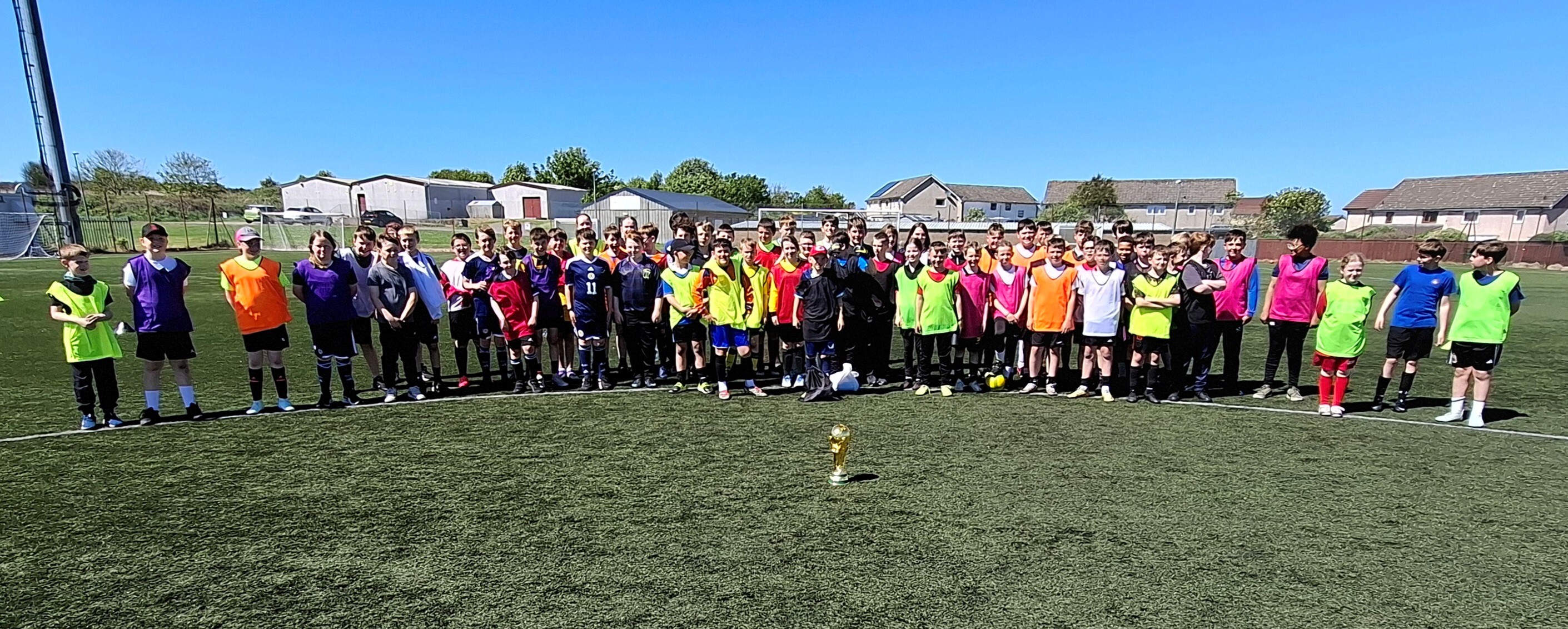 A large group of primary school pupils in colourful bibs standing on an artificial football pitch in sunshine, with a World Cup trophy in the foreground