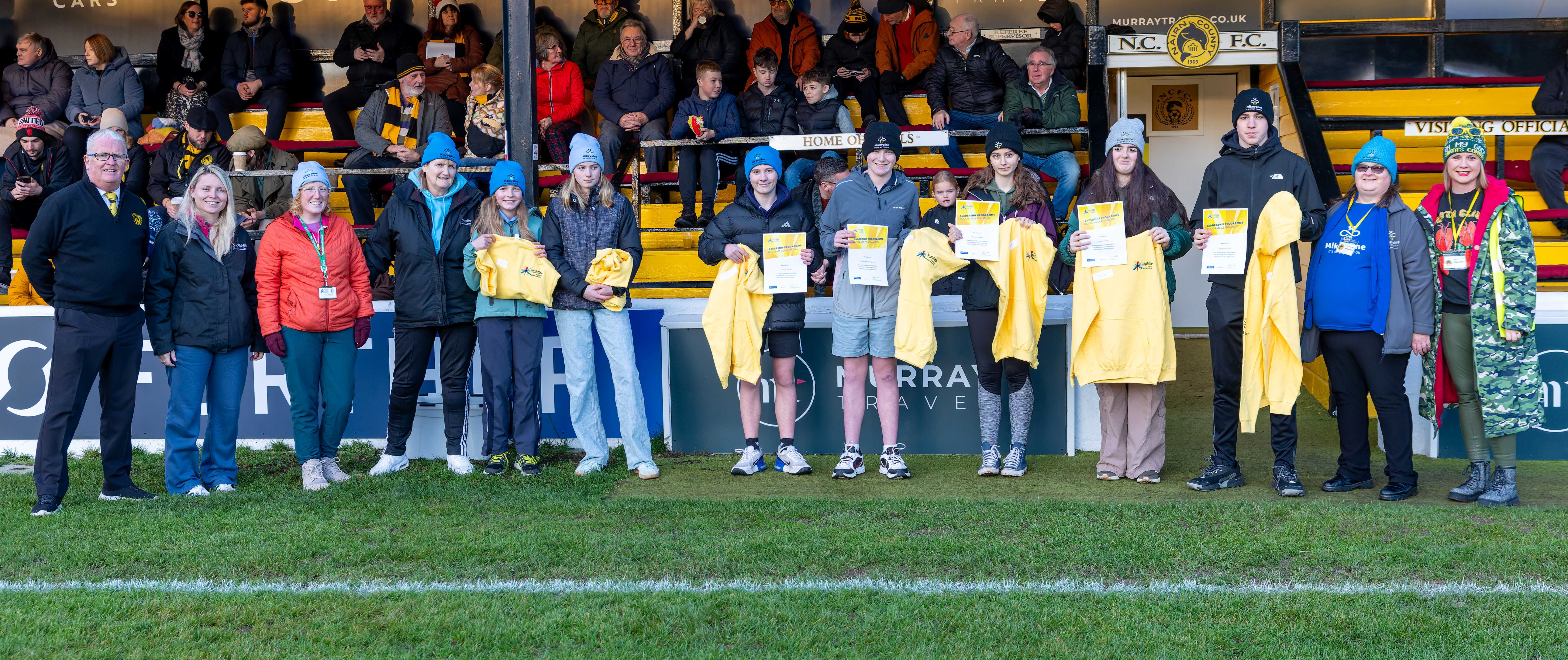 13 people standing in front of a football stand. Among them are seven young people holding up yellow hooded jumpers.