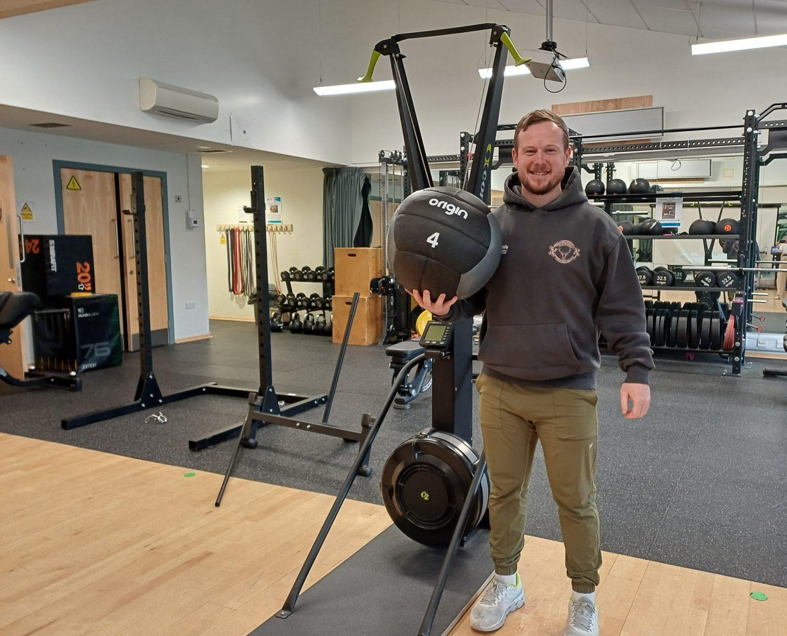 A fitness instructor standing in a gym holding a black medicine ball labeled "origin 4," surrounded by gym equipment.