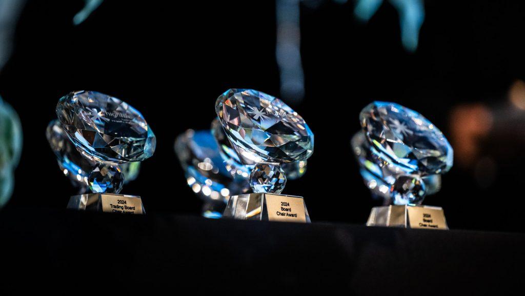 Three crystal awards with diamond-shaped tops and gold plaques reading '2024 Trading Board,' '2024 Board Chair Award,' and '2024,' displayed on a black tablecloth against a dark background.