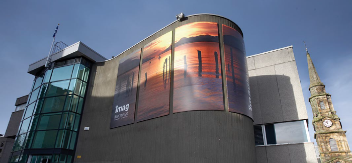A large building with a curved feature to the right and a glass frontage to the left. There is a church steeple in the background.