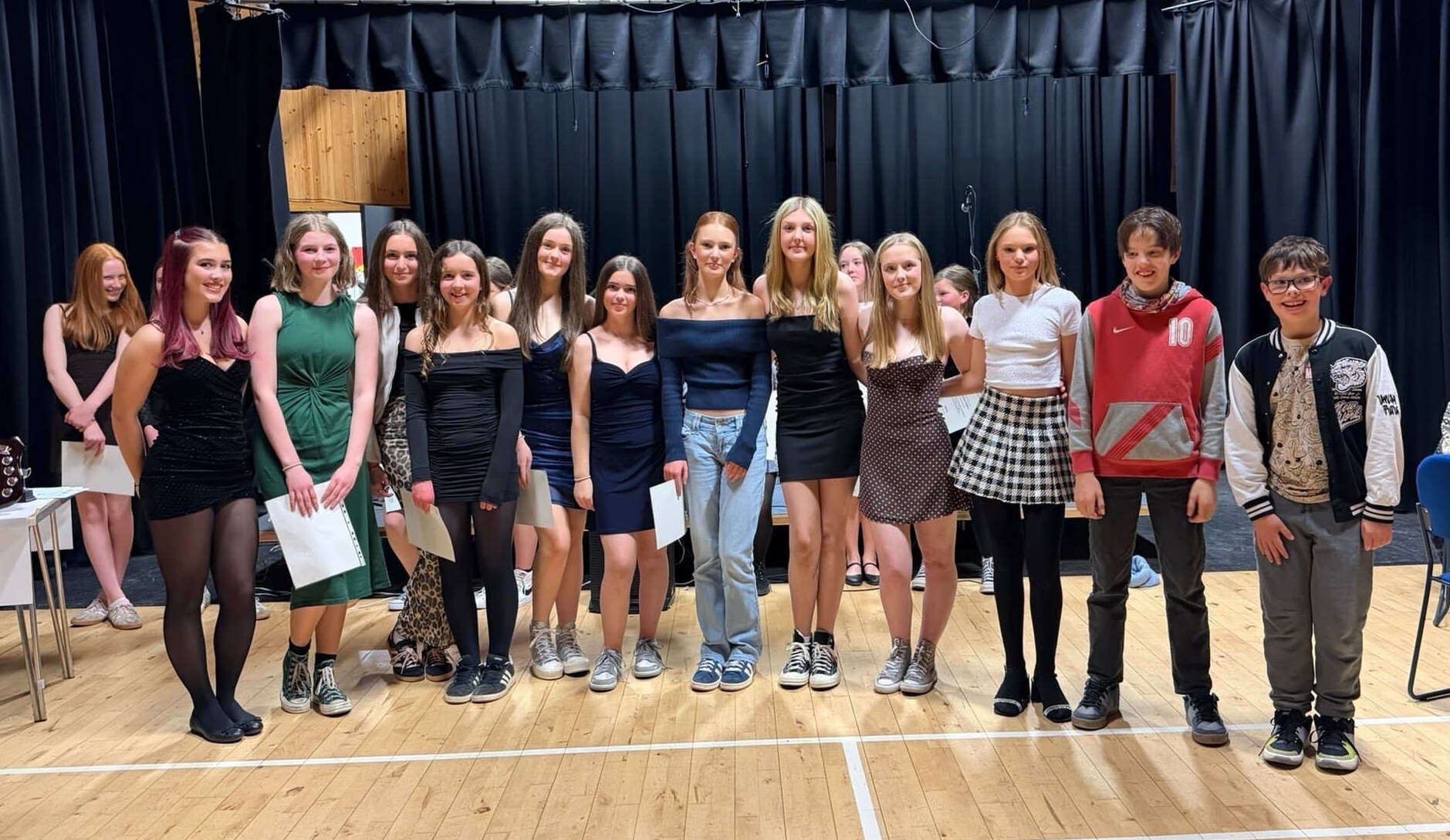 A group of 12 young people lined up in a sports hall with blue curtains behind.