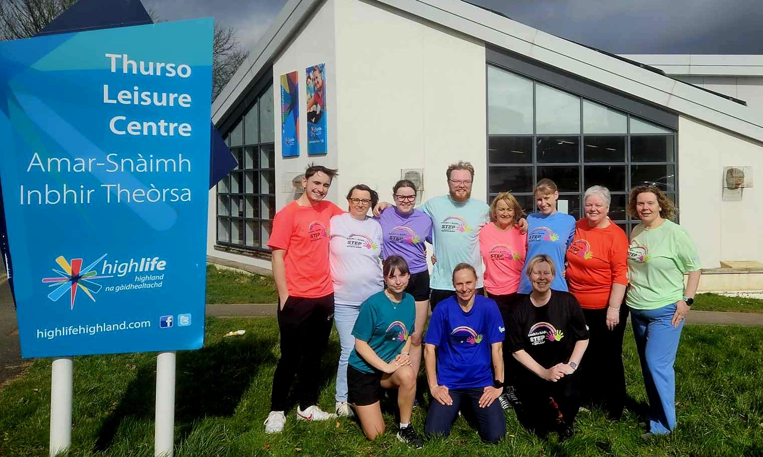 11 adults in a variety of coloured sportswear standing outside the Thurso Leisure Centre. There is a large blue Thurso Leisure Centre sign to the left of the group.