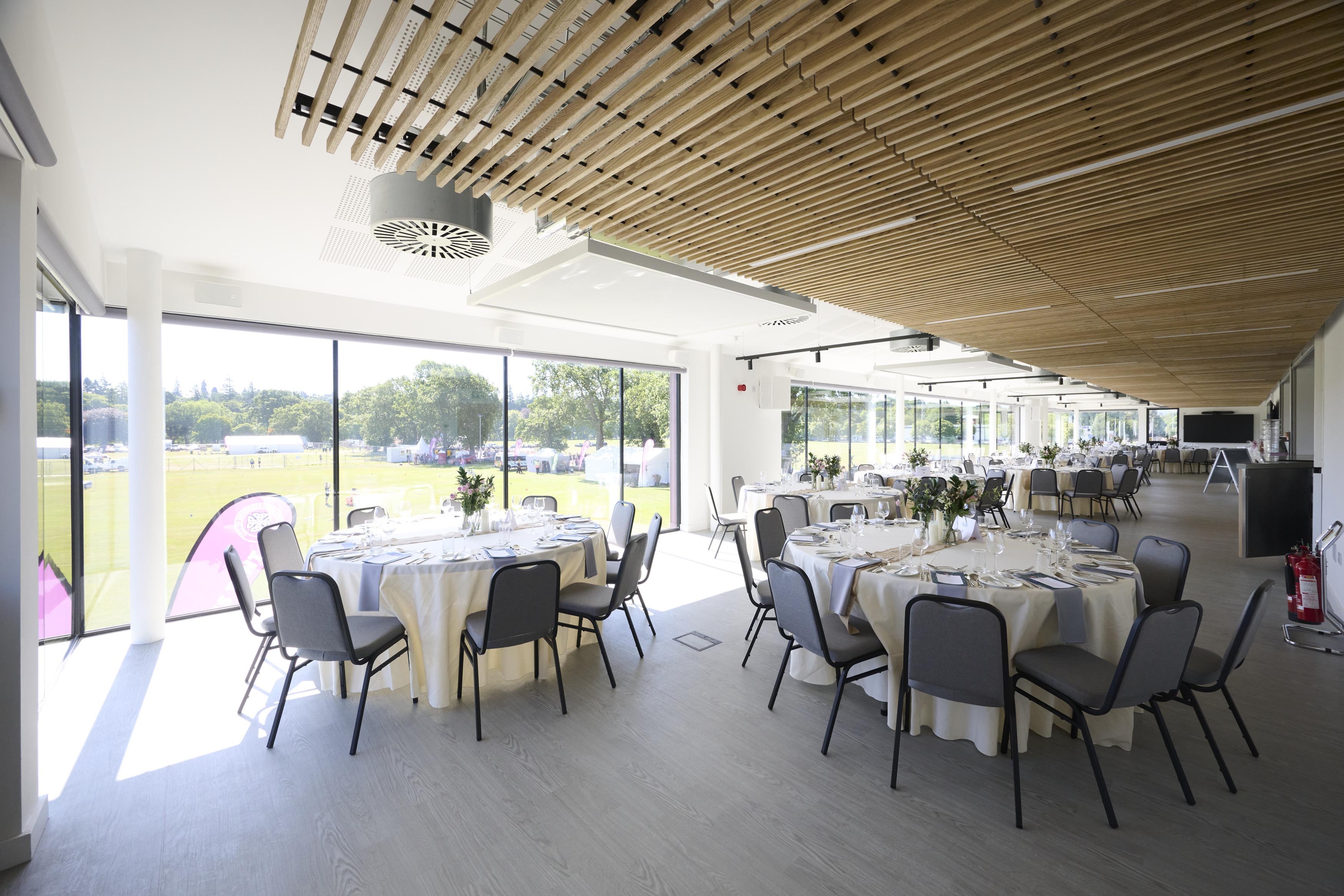 Bright, modern function room with round tables set for a meal, floor‑to‑ceiling windows, and a wooden slatted ceiling overlooking outdoor sports fields.