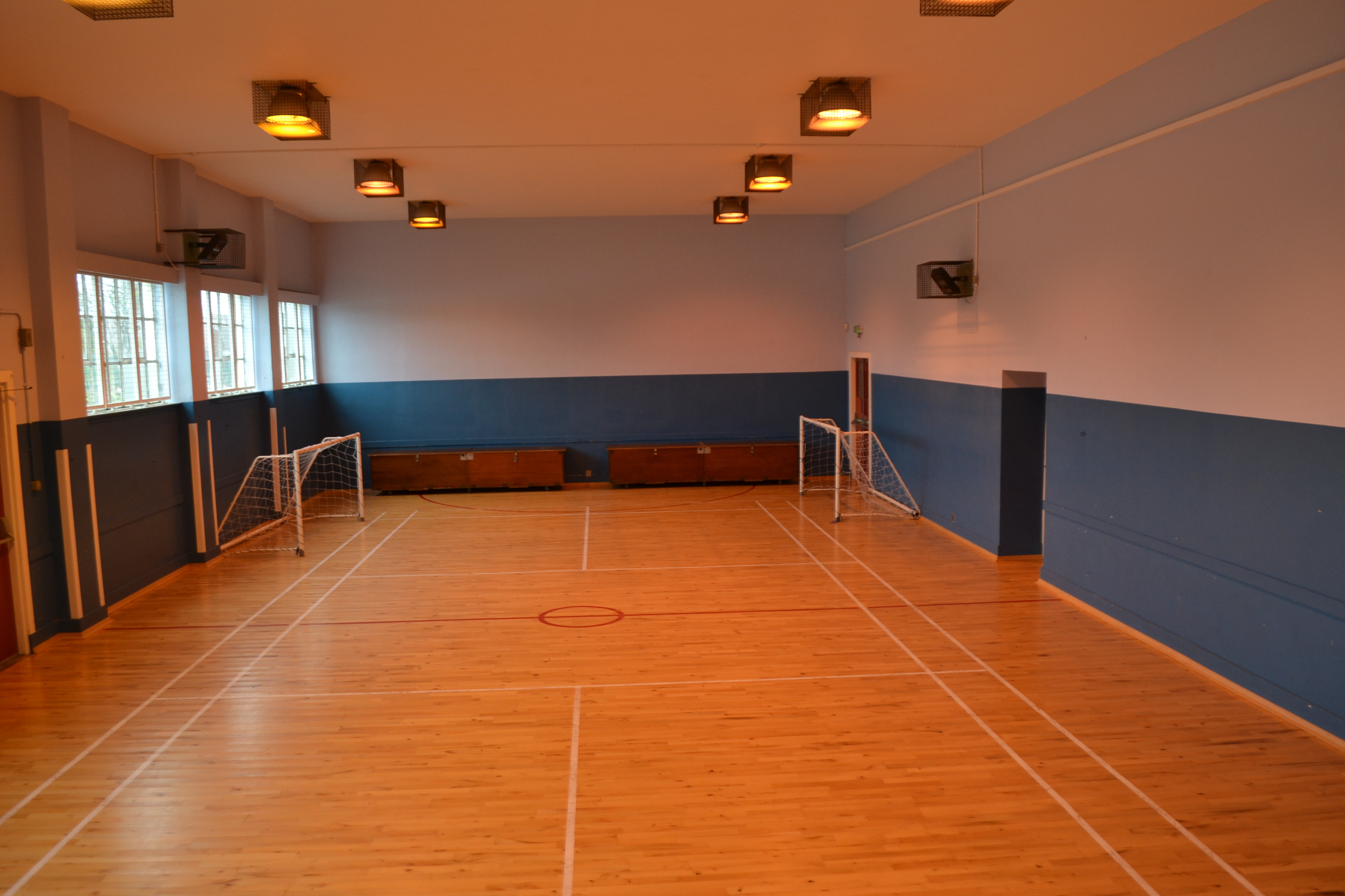 The gymnasium is a beautifully designed space, illuminated by natural light streaming in from windows on one side. It features a badminton court flooring, with two goals positioned at either end of the hall.