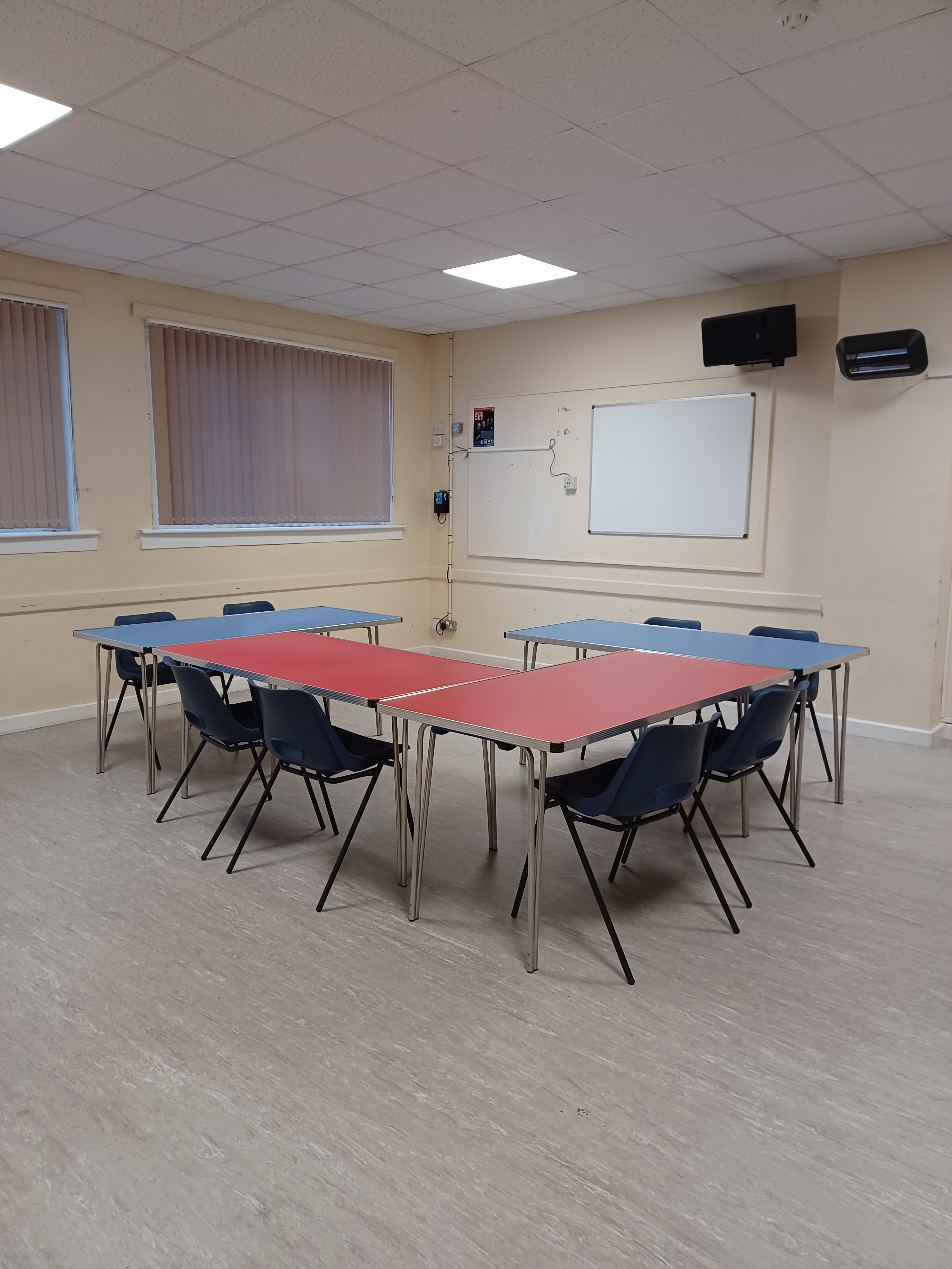 The room is bright and spacious, featuring linoleum flooring and a white board on the wall. Tables and chairs are arranged in the centre of the room.
