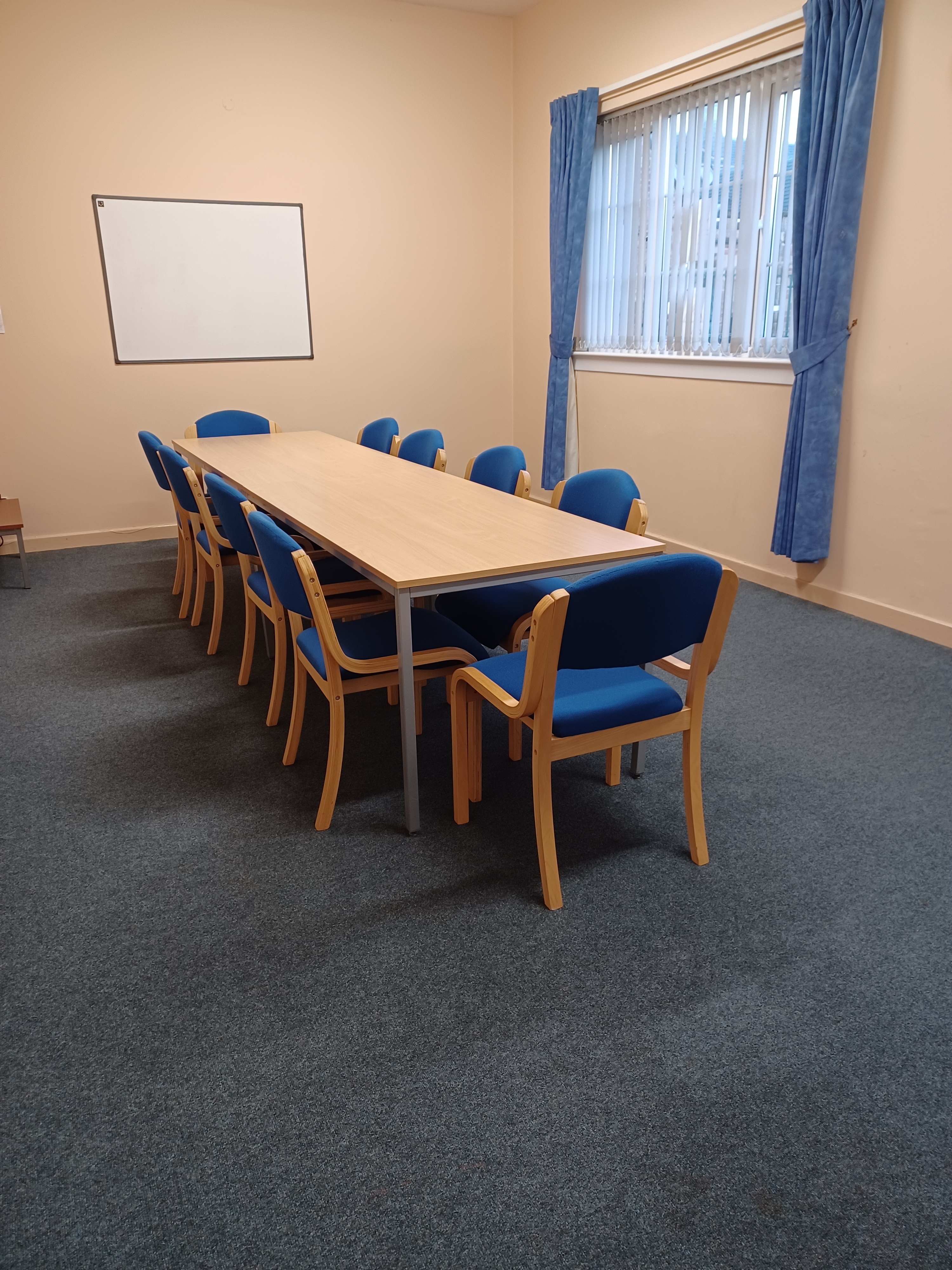 The room is elongated and features natural light streaming in from a curtained window on one side. It is furnished with a long meeting table, surrounded by chairs and a white board on the wall.