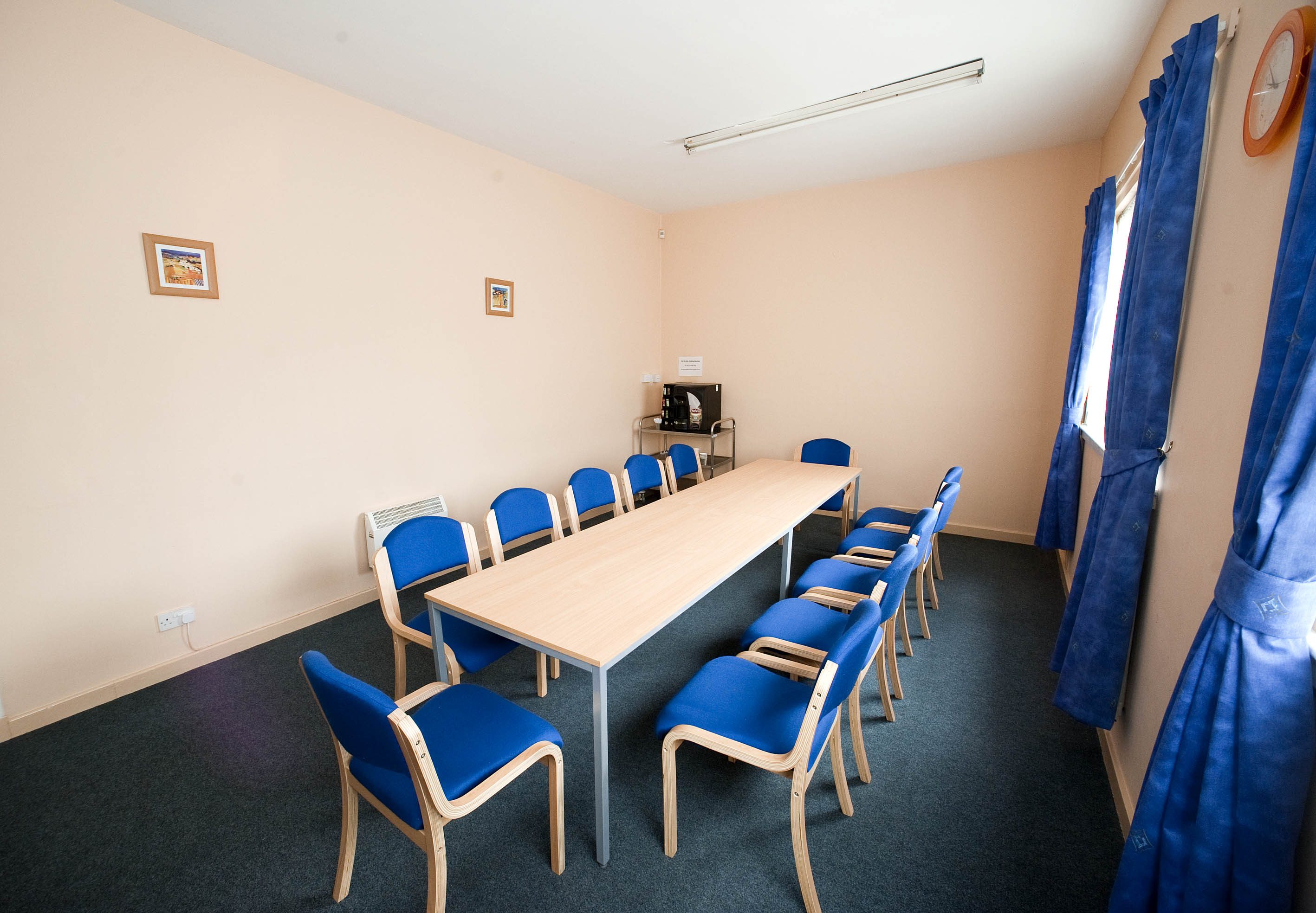 The room is elongated and features natural light streaming in from two curtained windows on one side. It is furnished with a long meeting table, surrounded by chairs, creating an inviting space for discussions and collaboration.