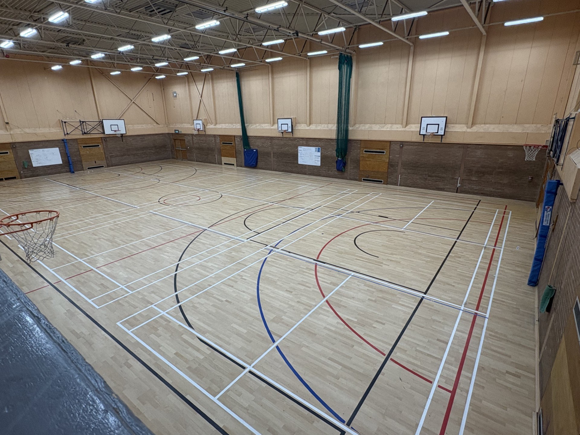 A spacious games hall featuring four badminton courts, complete with basketball floor markings and multiple basketball hoops positioned on all four walls.