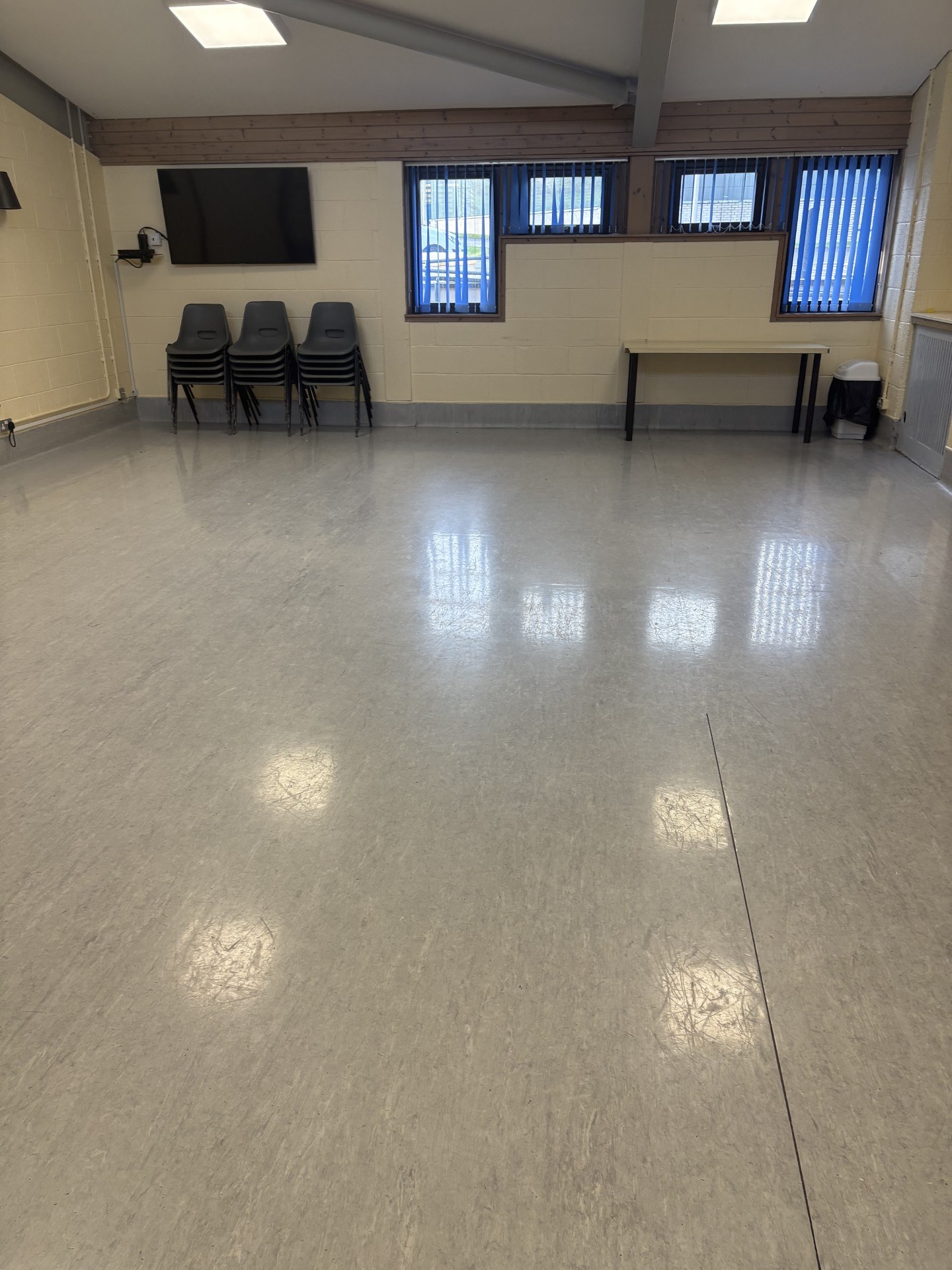 The Graham Ross suite 1 features laminated flooring and stone walls, enhanced by natural lighting from the windows. It is equipped with tables, stacked chairs, and a wall-mounted television screen.