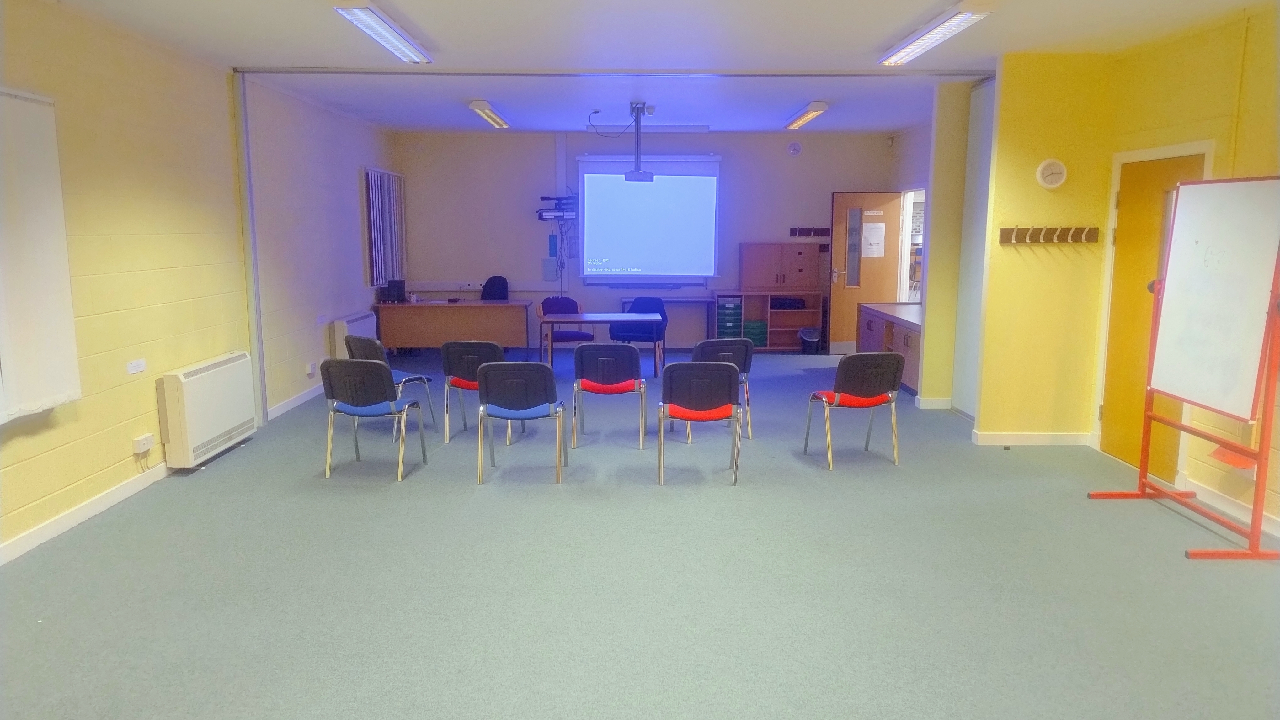Spacious room featuring a white presentation screen mounted on the far wall. Chairs are arranged to face the screen, and an additional whiteboard is positioned against the side wall.