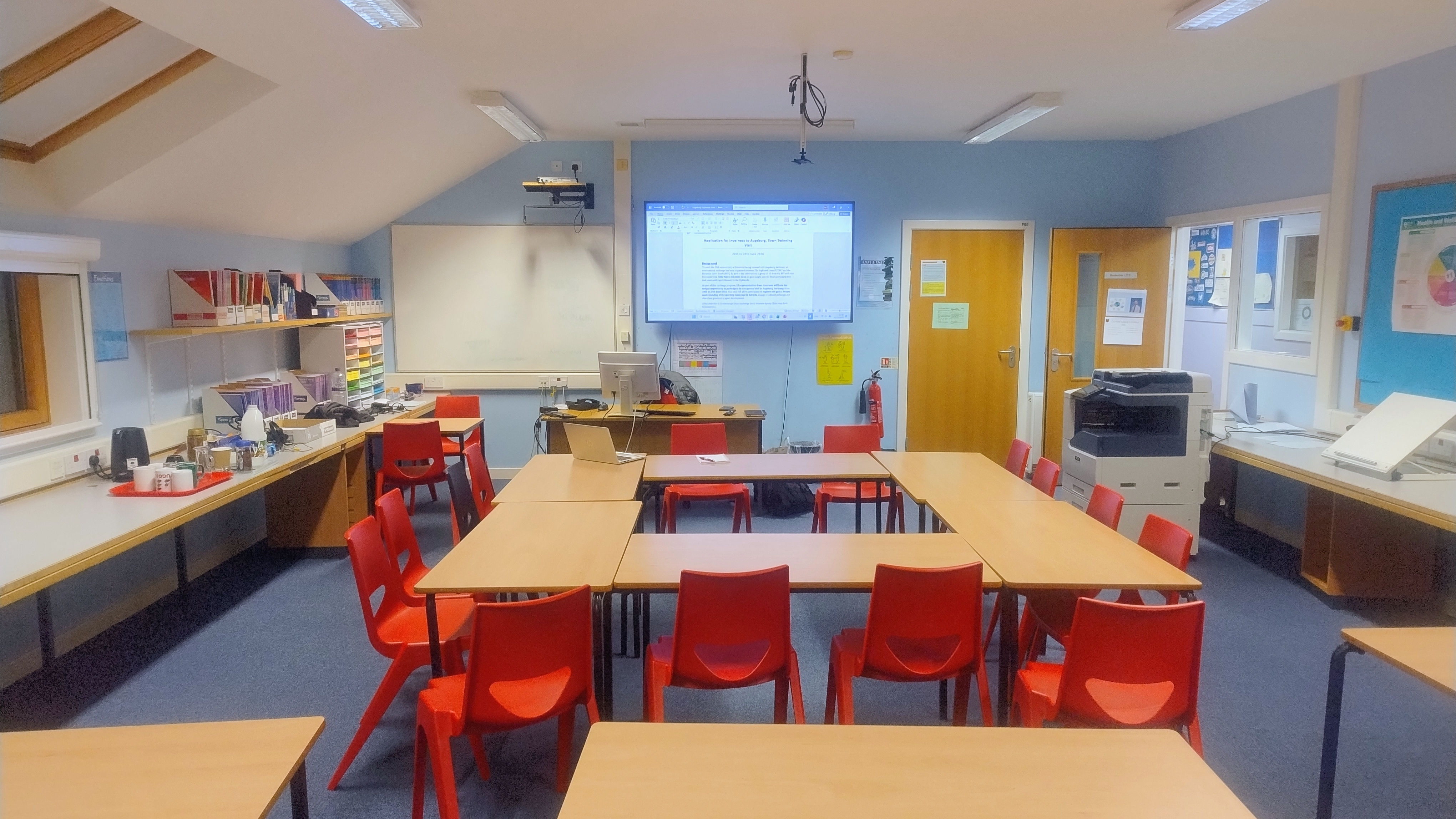 Classroom arranged with tables and chairs configured in a square formation at the center of the room. Additional tables are positioned at the back and front of the room. There is a large wall-mounted TV screen, accompanied by an extra whiteboard.